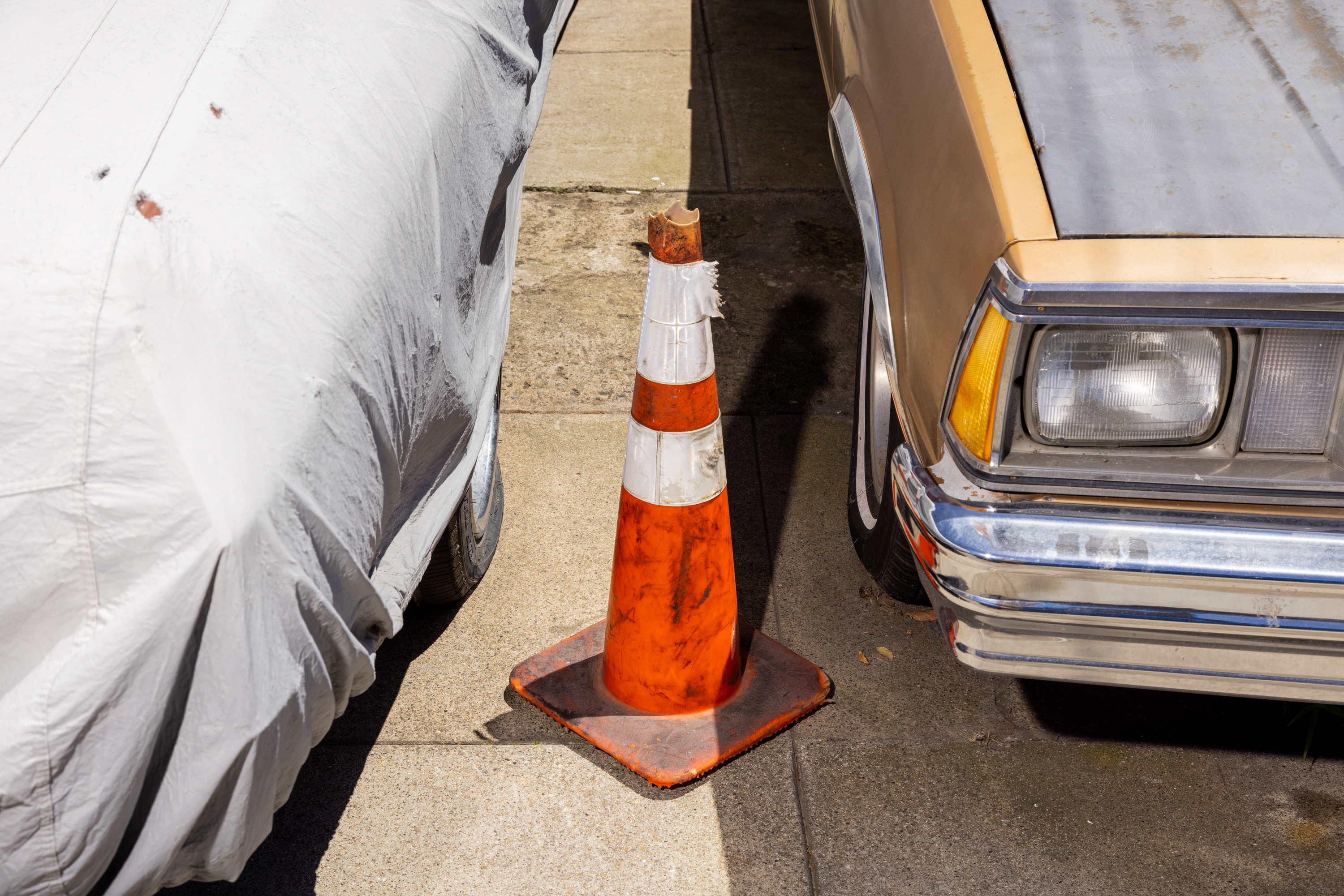 A traffic cone sits between two parked cars, one covered with a tarp and the other exposed.