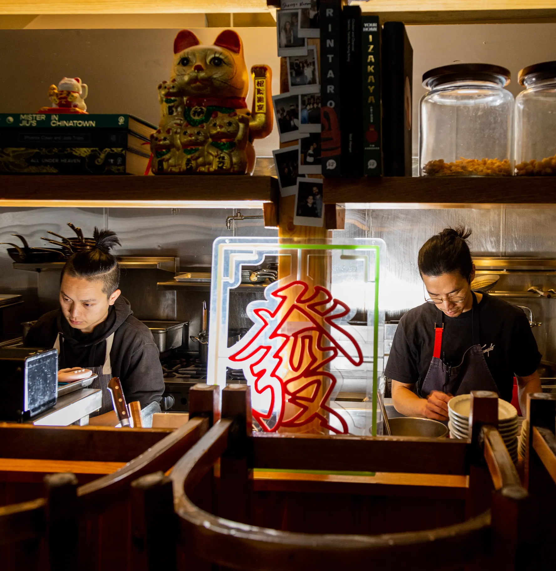 Two chefs work in a kitchen with Asian decor, including a maneki-neko (lucky cat) and a neon sign.