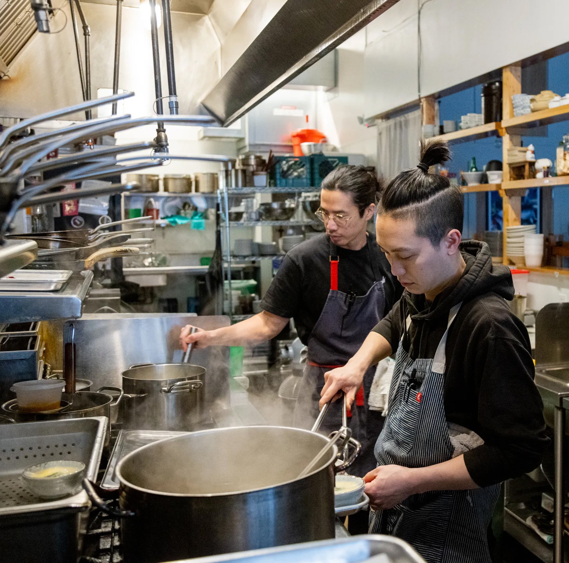 Two chefs are working in a busy commercial kitchen with pots steaming on the stove.
