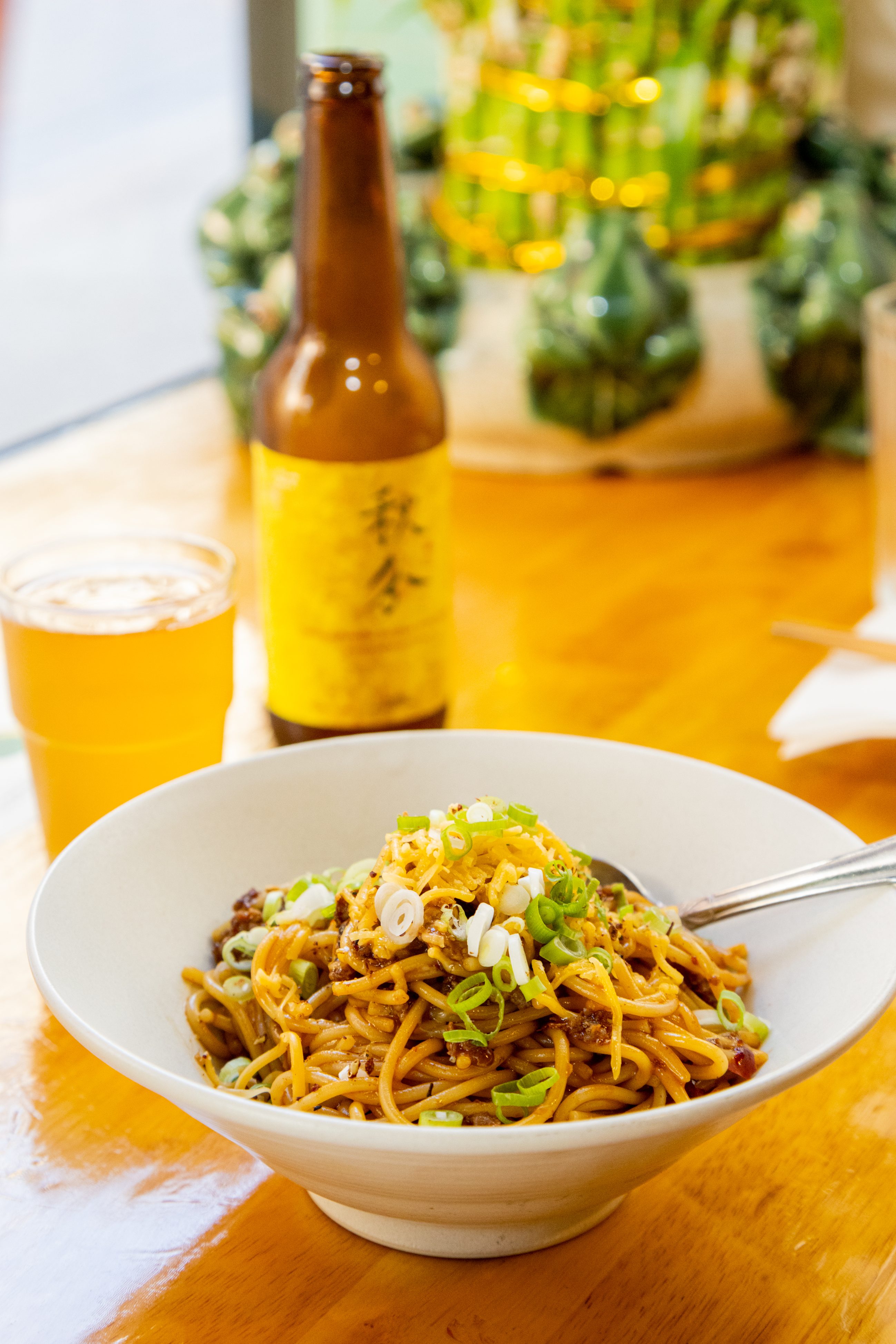 A bowl of noodles garnished with green onions beside a beer bottle and glass. Blurred plants in the background.