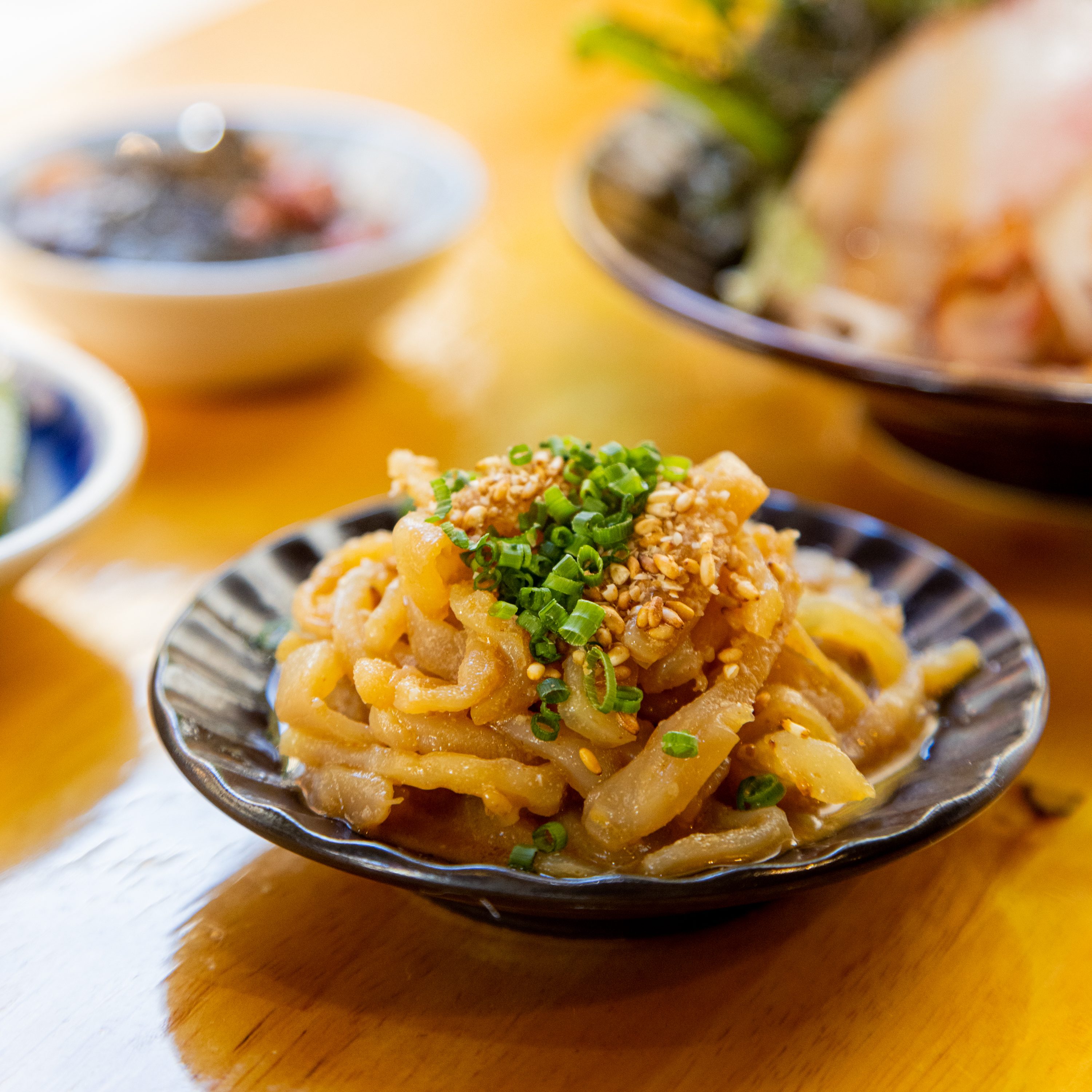 A plate of udon noodles topped with green onions and sesame seeds, with blurry bowls in the background.