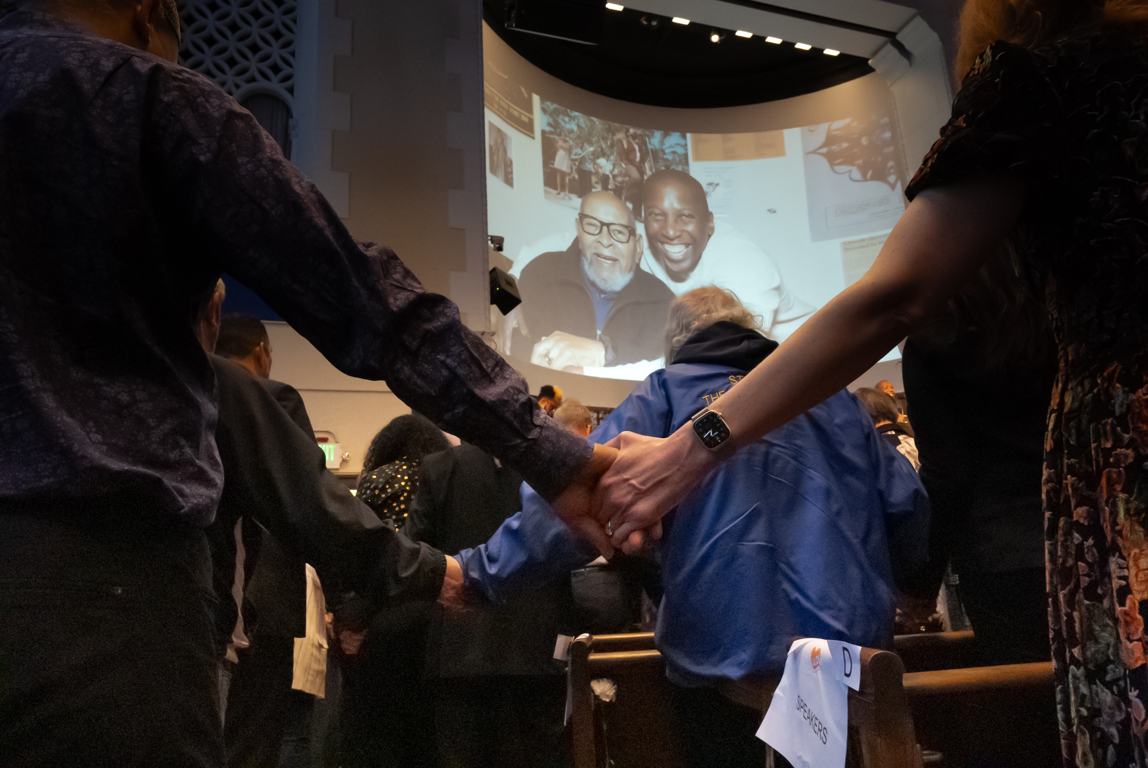 People holding hands at a service with a screen showing two smiling men.