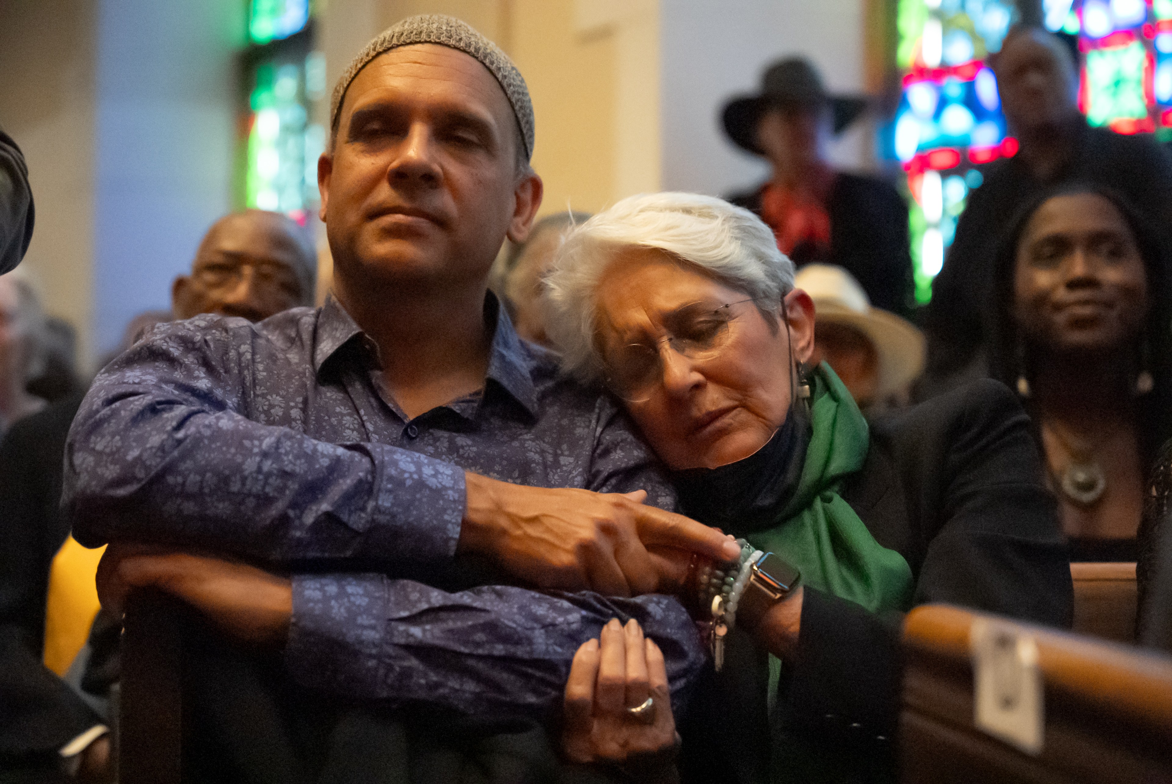 A man and woman in a church, woman leaning on man's shoulder, both appear contemplative, stained glass and other attendees in background.
