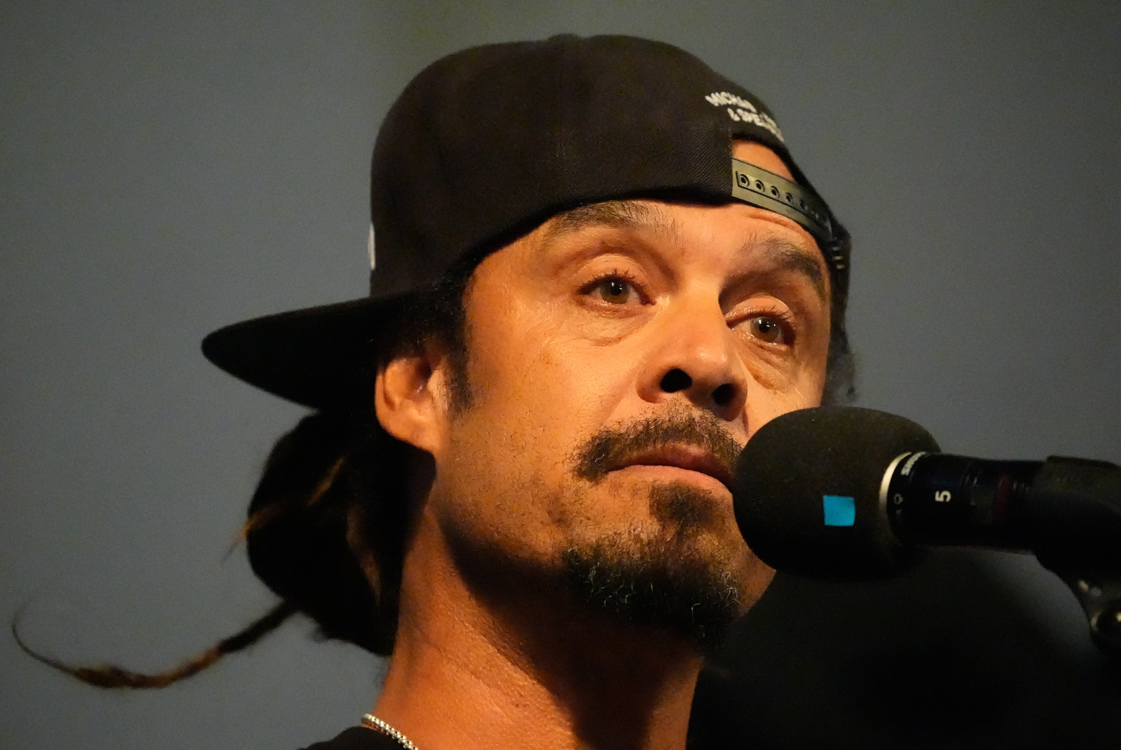 A man speaks into a microphone, wearing a cap, with a solemn expression and a thin mustache.