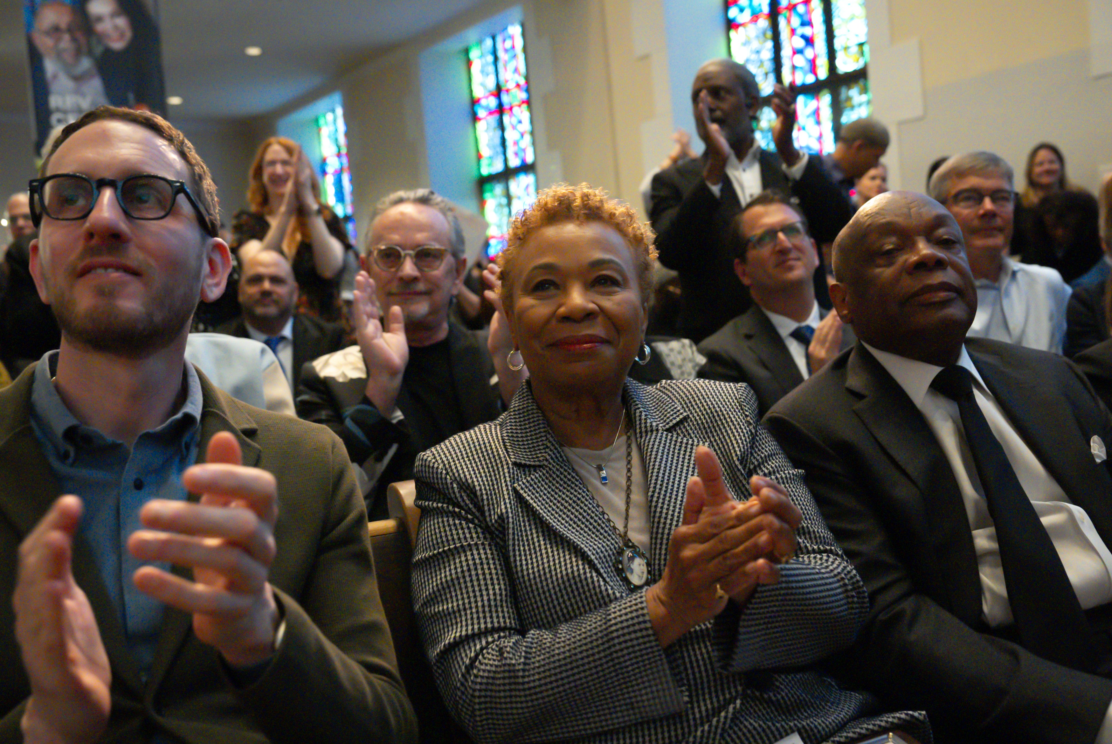A diverse crowd clapping indoors, some smiling, with stained glass windows in the background.