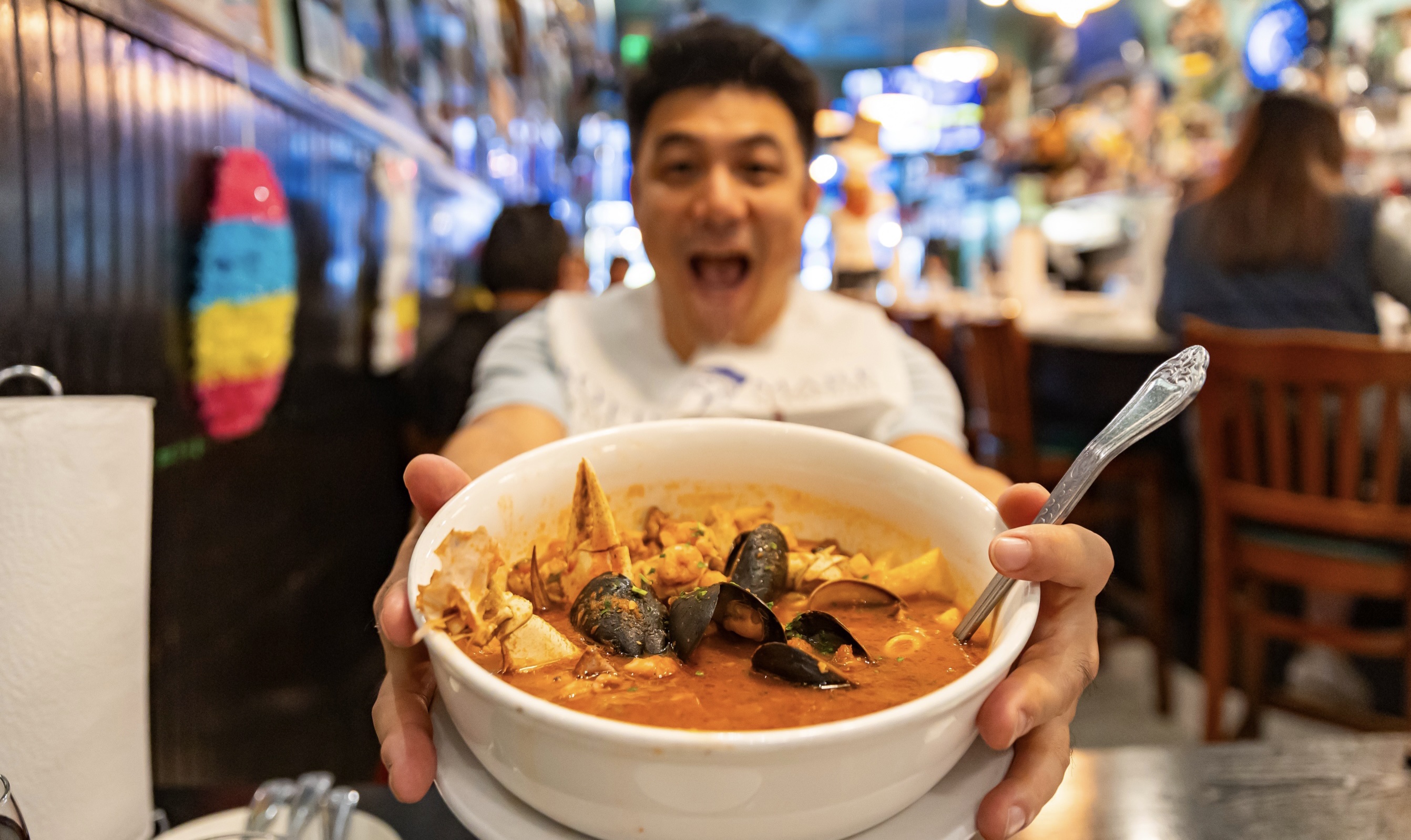 A man excitedly presents a large bowl of seafood soup, prominently displaying mussels and rich broth, in a vibrant, decorated restaurant.