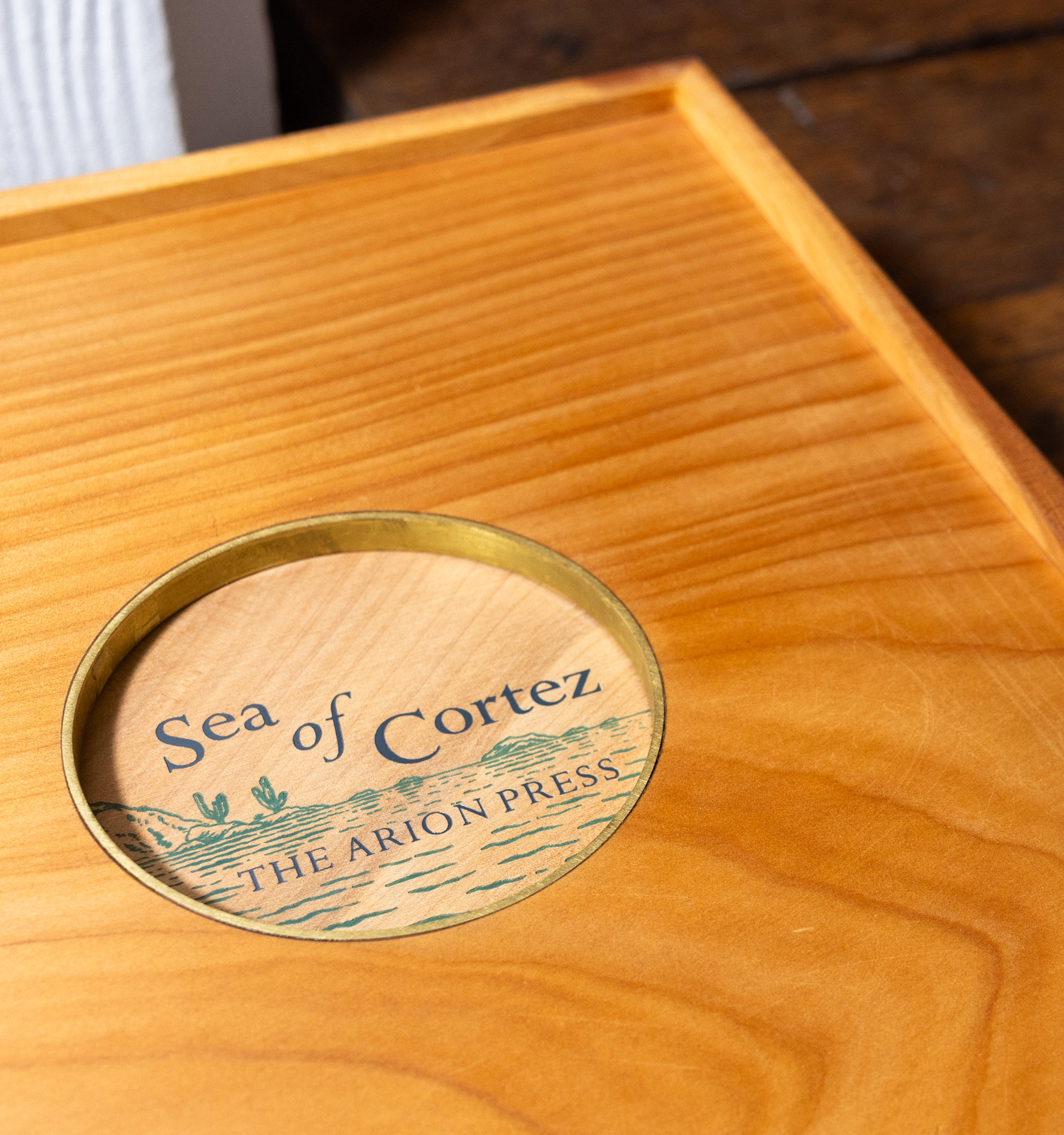A wooden lid features an inset, rounded, brass-bound window displaying text &quot;Sea of Cortez - The Arion Press&quot; over a stylized wave and cactus illustration.