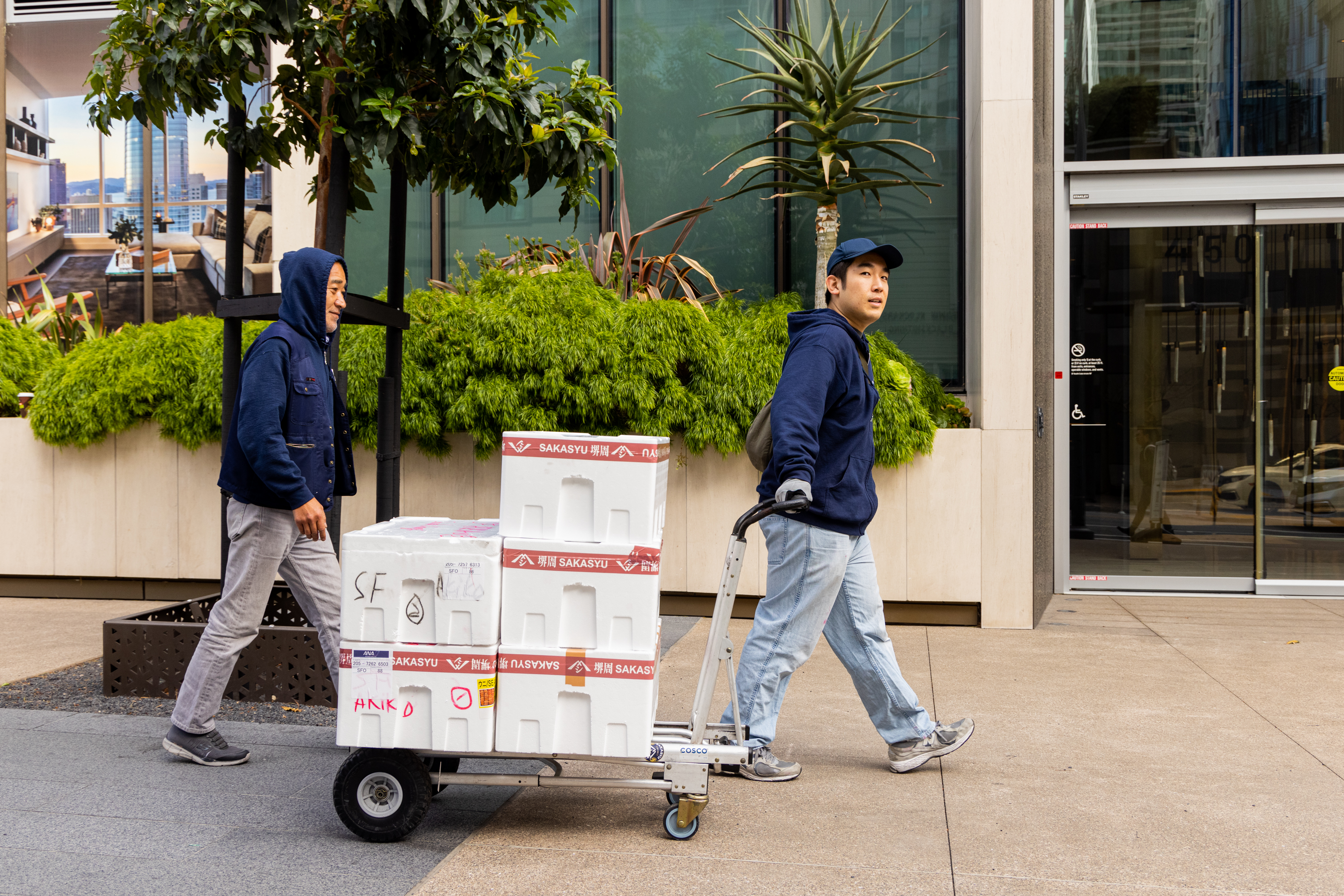 Two men are walking on a sidewalk, one pulling a handcart loaded with white boxes. They are dressed in casual clothing, and the background features greenery and modern buildings.