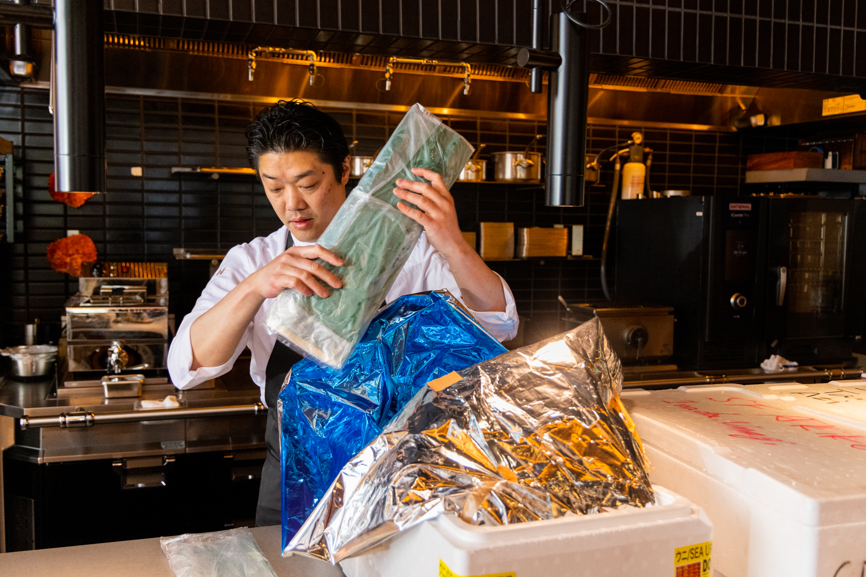 A chef in a white uniform stands in a kitchen, unwrapping items from shiny foil and plastic bags, possibly unpacking supplies from large styrofoam boxes.