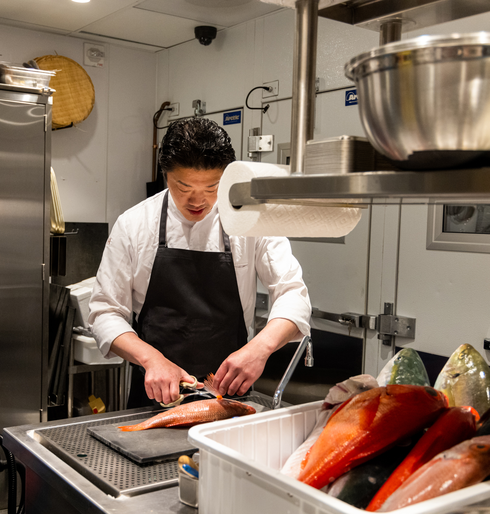 A chef wearing a white coat and black apron is filleting a fish in a professional kitchen. Several whole fish are in a white tub beside him, with utensils nearby.