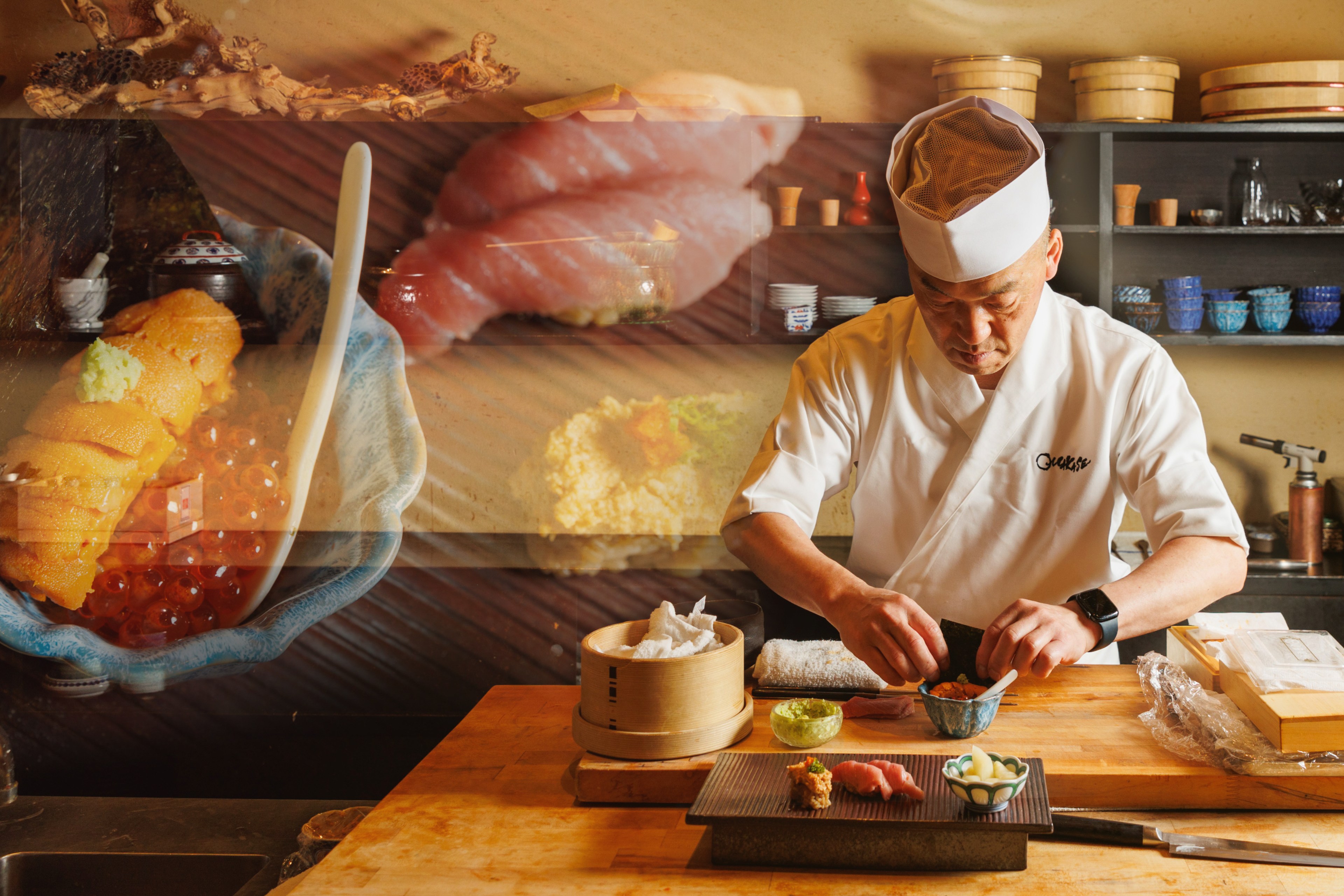 A sushi chef, dressed in white, prepares an intricate dish at a wooden counter with sushi and various ingredients. The background shows bowls and kitchen shelves.