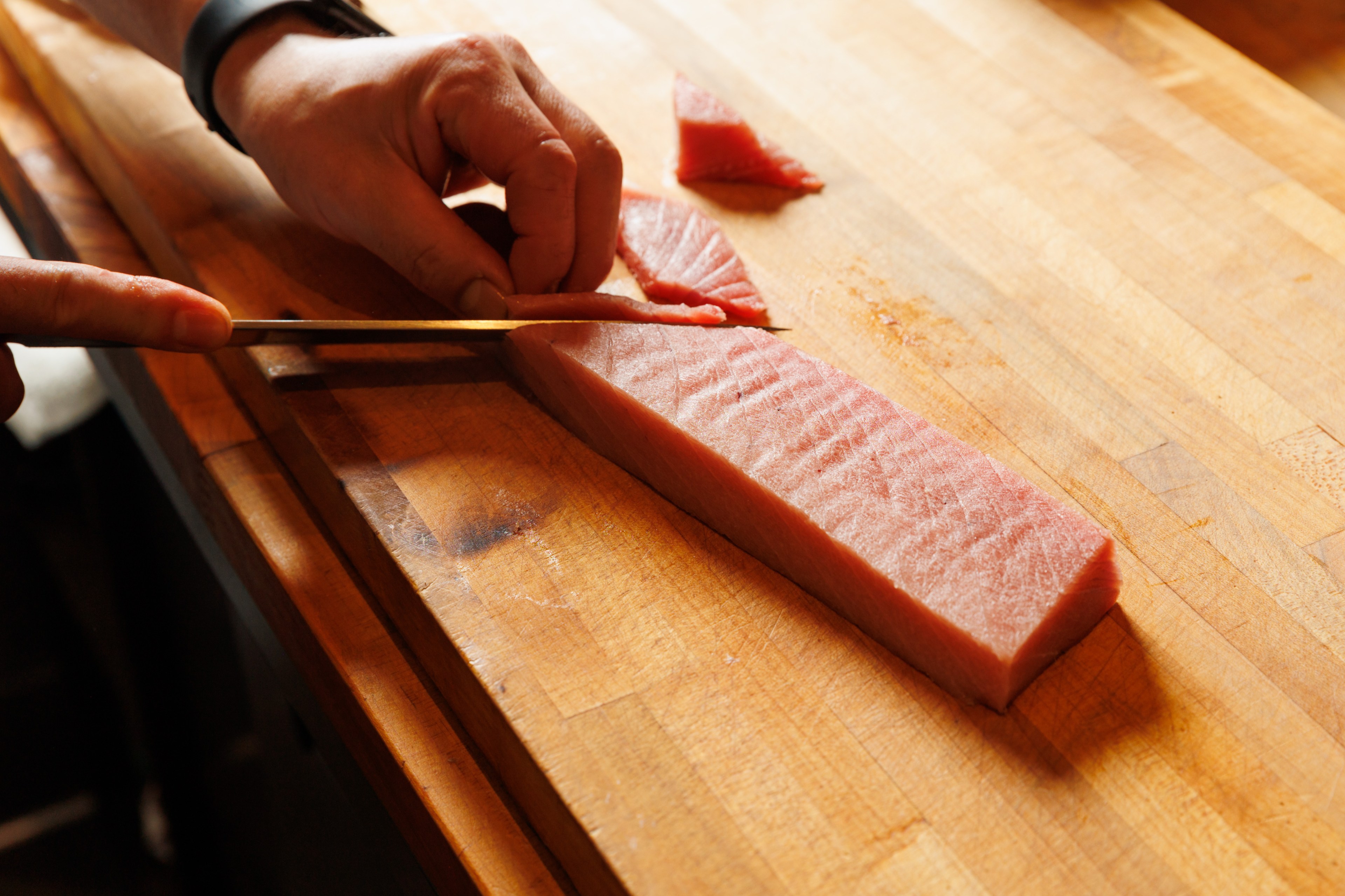 A person uses chopsticks to delicately slice raw tuna on a wooden cutting board. The tuna has a firm, pink texture, and a piece has already been cut.