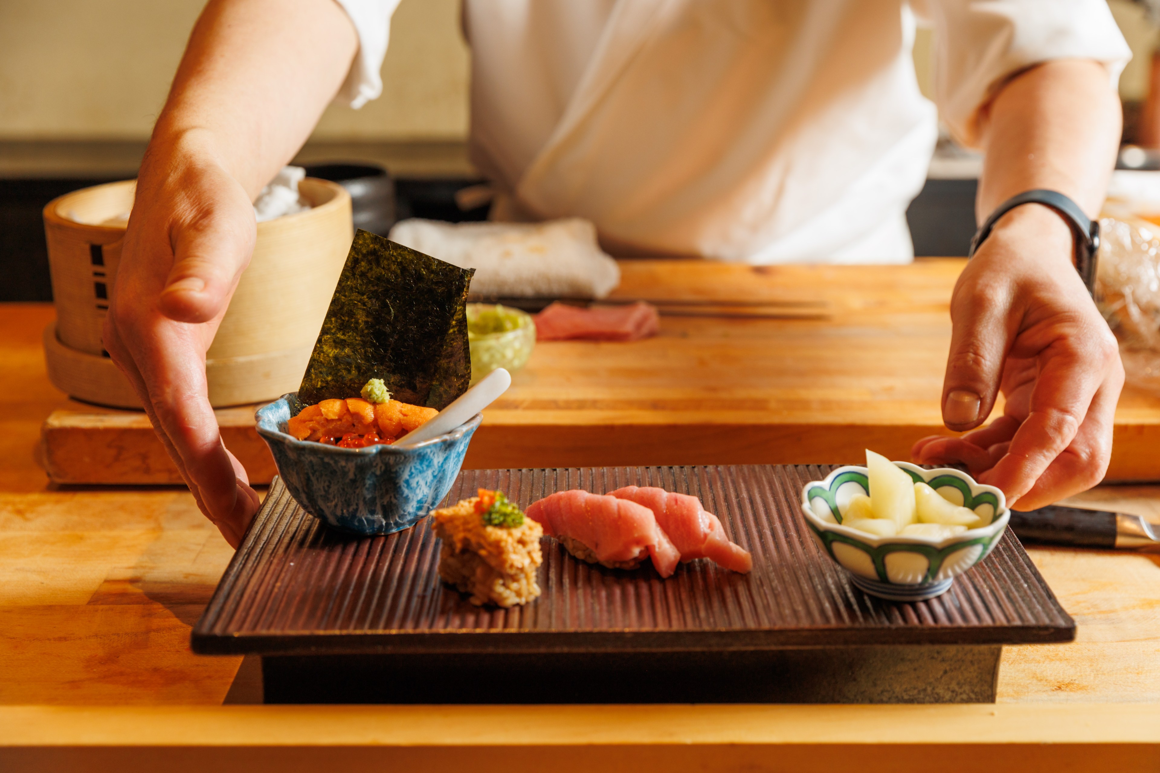 A chef presents a wooden platter with assorted sushi, including a blue bowl with seaweed, a small portion of sliced pink fish, and a white bowl with pickled vegetables.