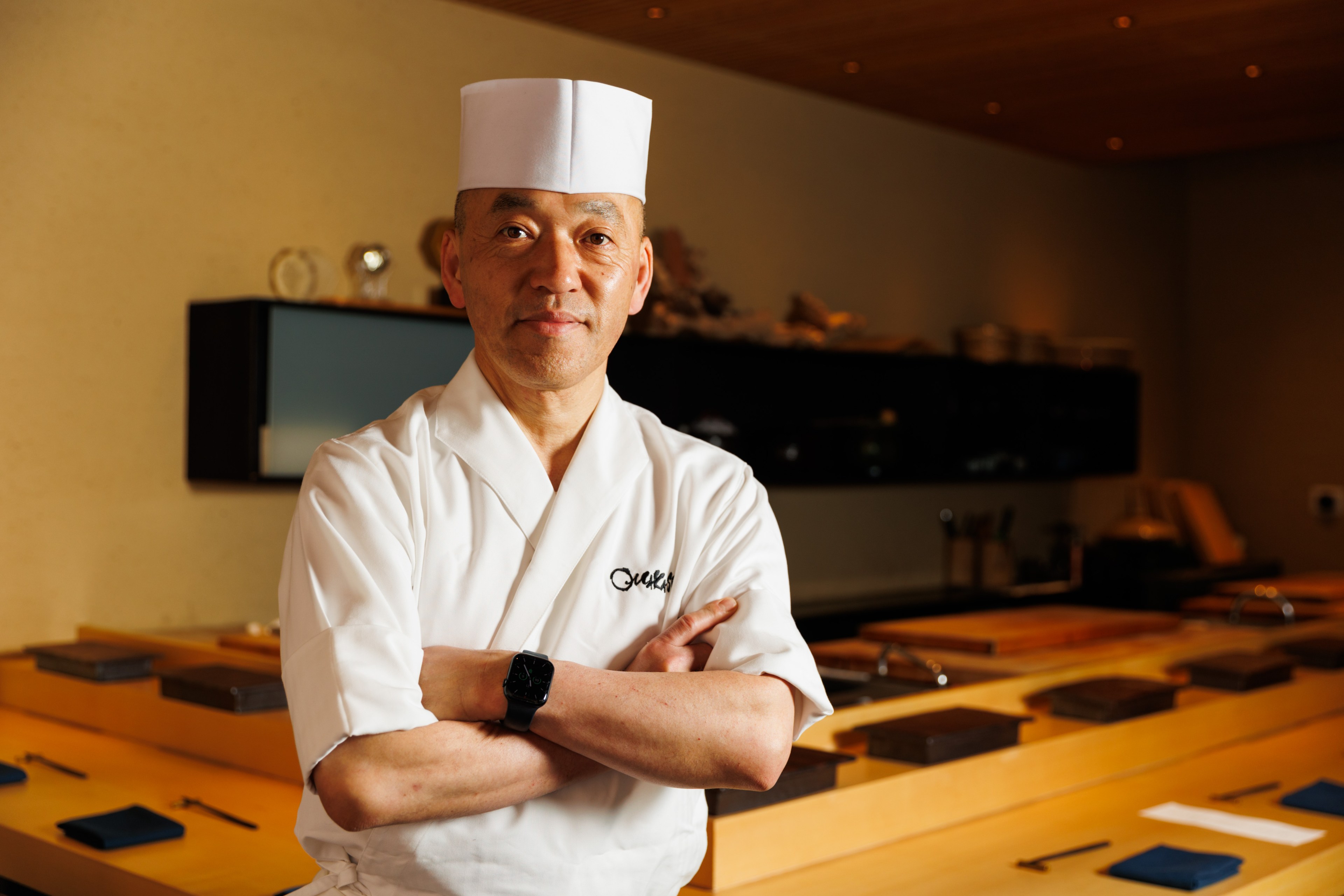 A chef in a white uniform and tall hat stands confidently with arms crossed in a warmly-lit, minimalist kitchen, with wooden countertops and shelves in the background.