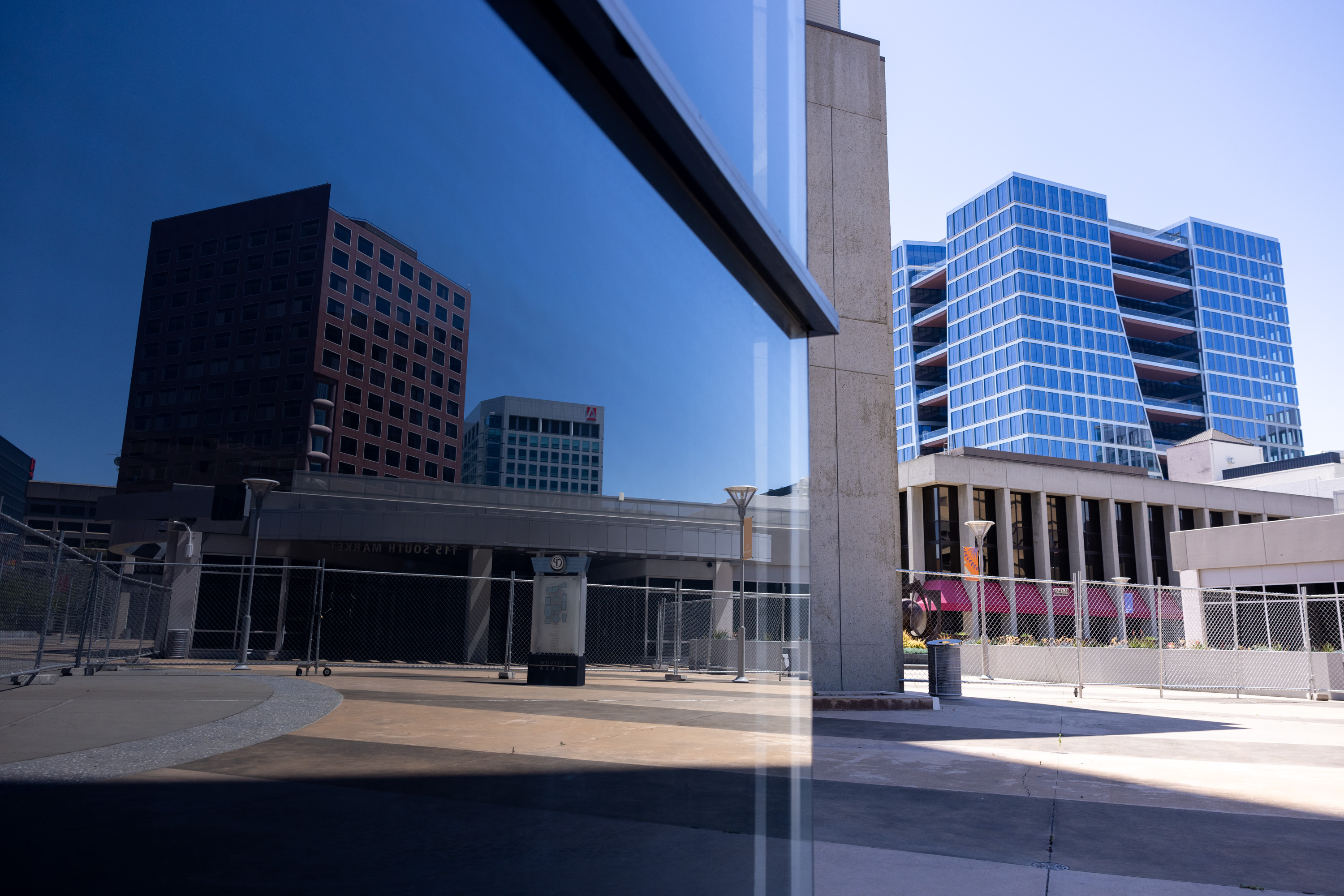 The image shows modern high-rise buildings with mirrored reflections, chain-link fences, and a clean, open concrete plaza under a clear blue sky.