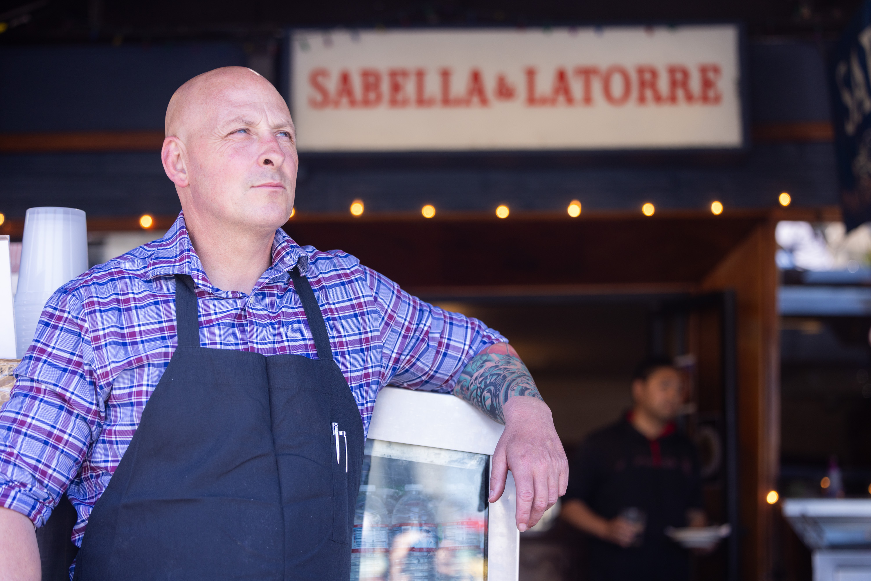 A bald man in a plaid shirt and black apron looks into the distance while leaning on a counter. Behind him, a &quot;SABELLA &amp; LATORRE&quot; sign is visible.