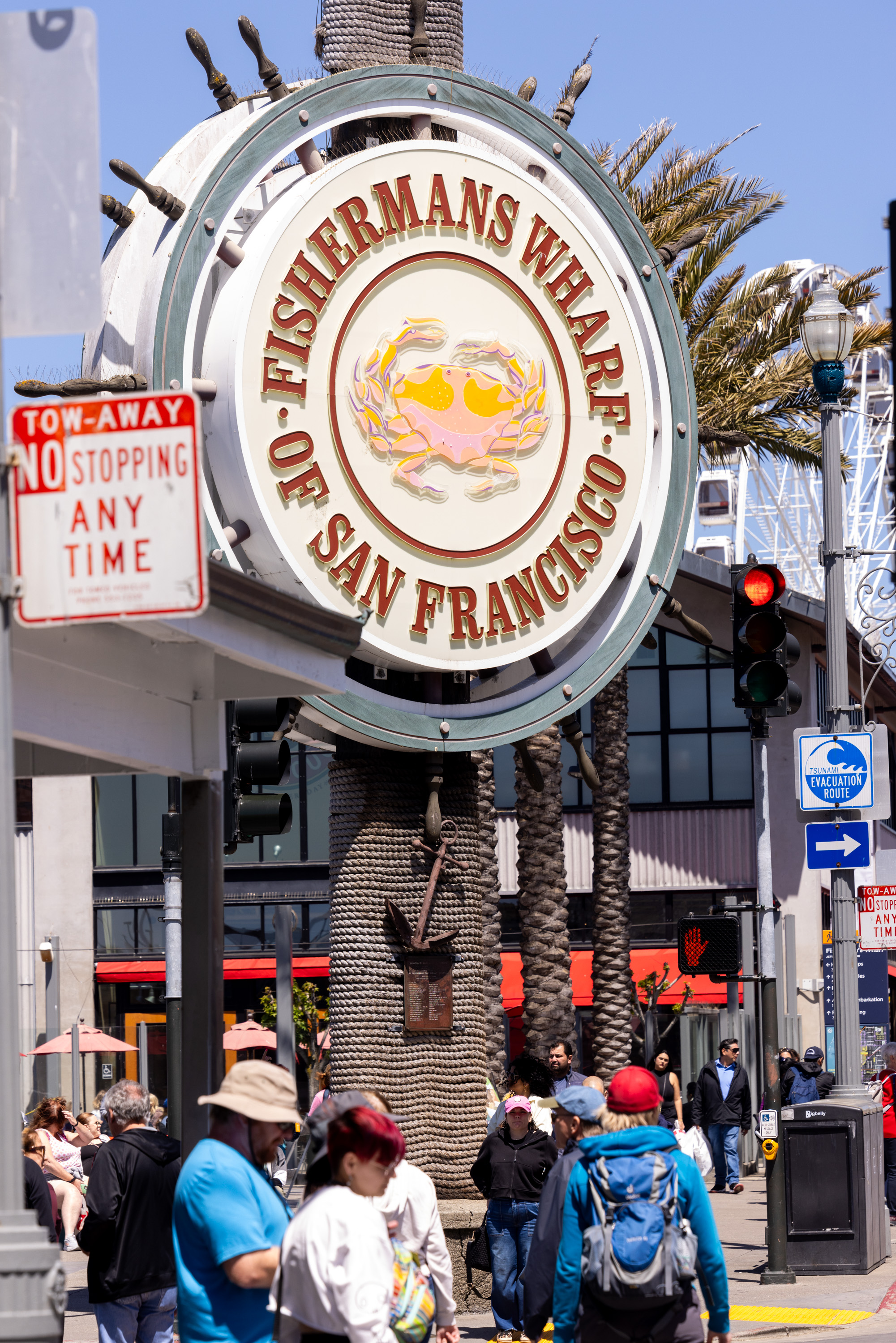 The image shows a busy street scene at Fisherman's Wharf, San Francisco, with a large circular sign and a crowd of people walking beneath it. Nearby, there's a &quot;No Stopping Any Time&quot; sign.