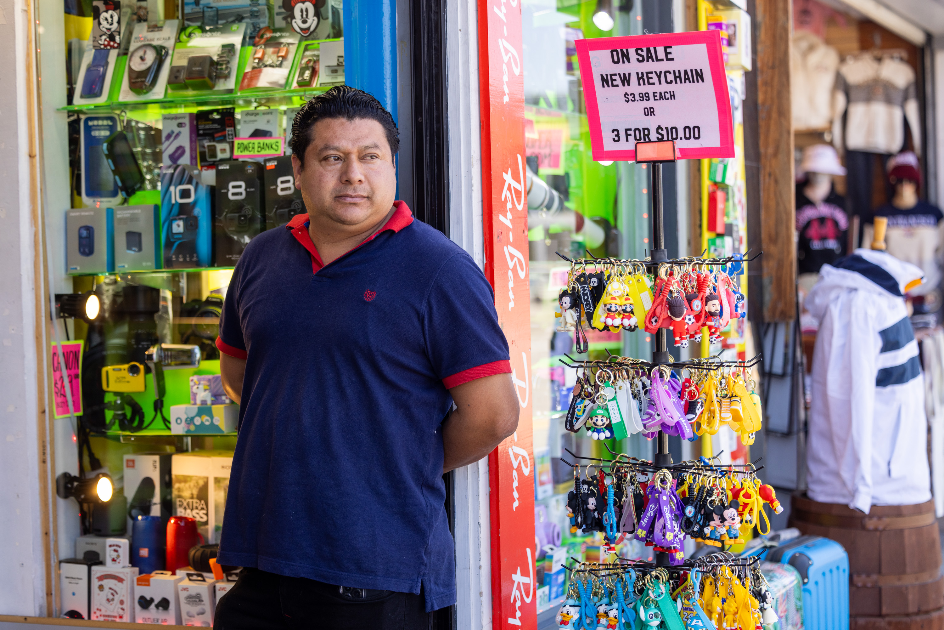 A man stands outside a shop filled with electronic gadgets and accessories, next to a display of colorful keychains with a sign reading "On Sale New Heychain $3.99 Each or 3 For $10.00."