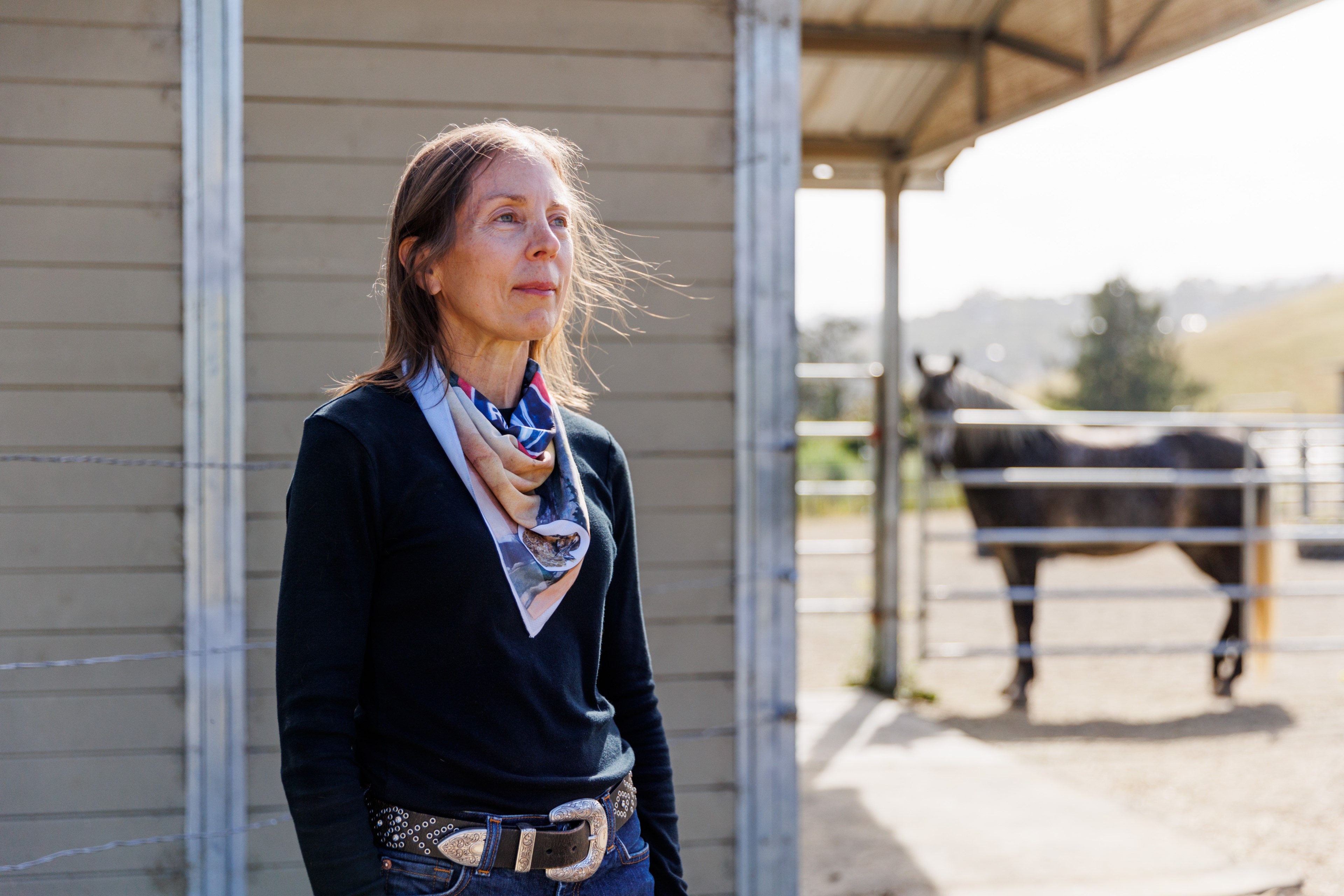 A woman with a colorful scarf and a black shirt stands outside near a timber building. A horse is visible in the background, enclosed in a fenced area.