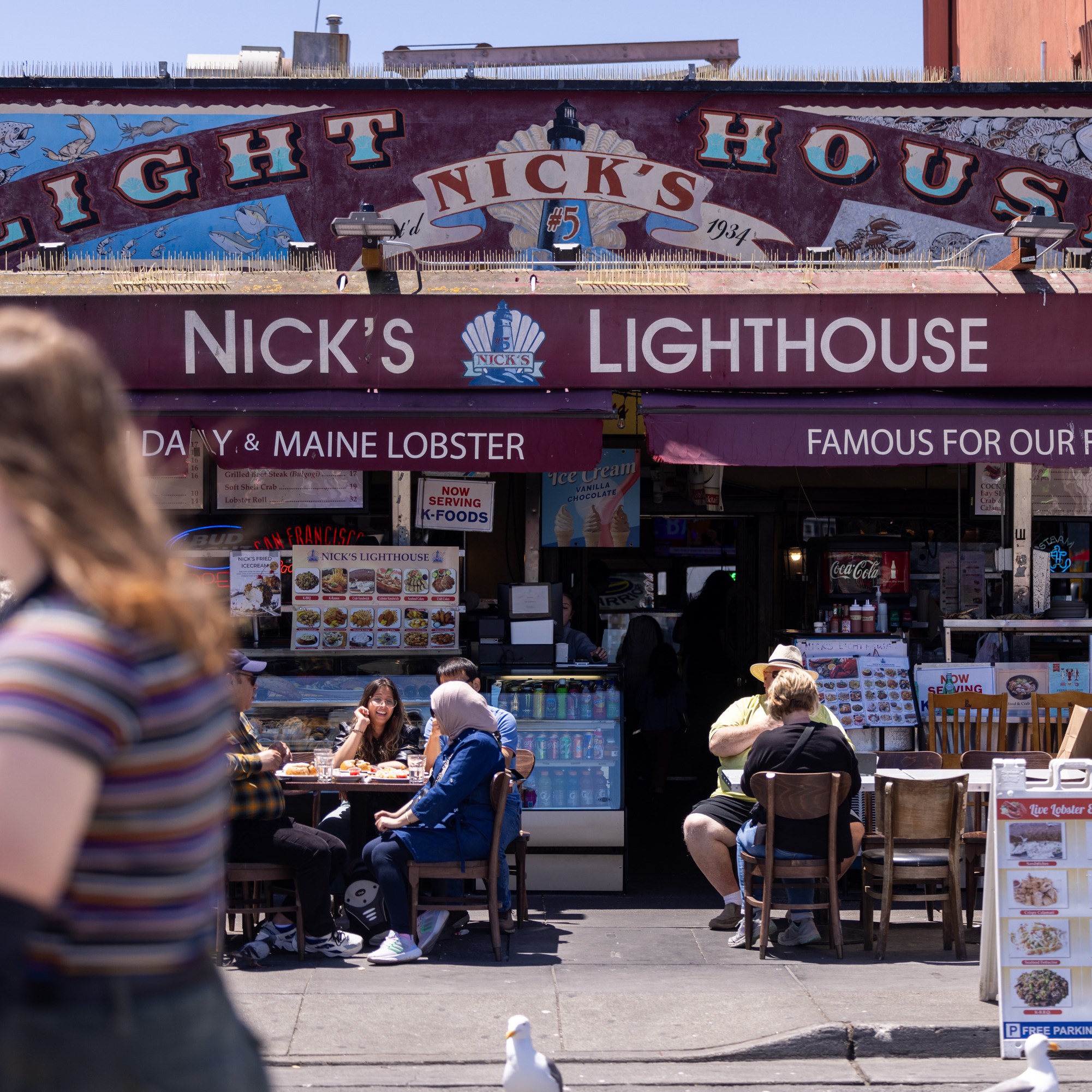 This image shows the exterior of Nick's Lighthouse restaurant, with its maroon canopy displaying its name. People are dining outside on a sunny day, enjoying seafood dishes.