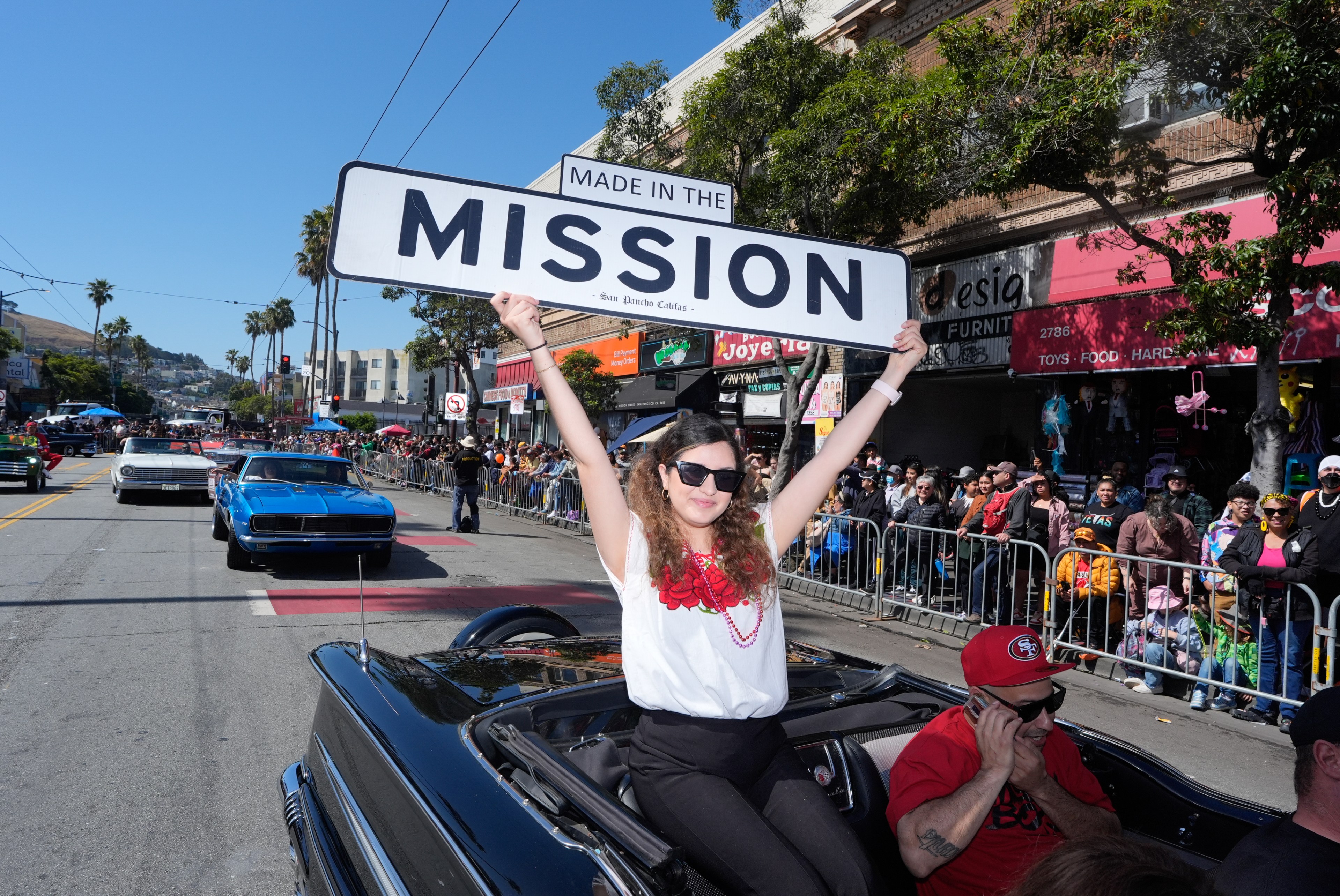 A woman in a red-embroidered white top and sunglasses holds a &quot;Made in the Mission&quot; sign, riding in a convertible during a parade with spectators lining the street.