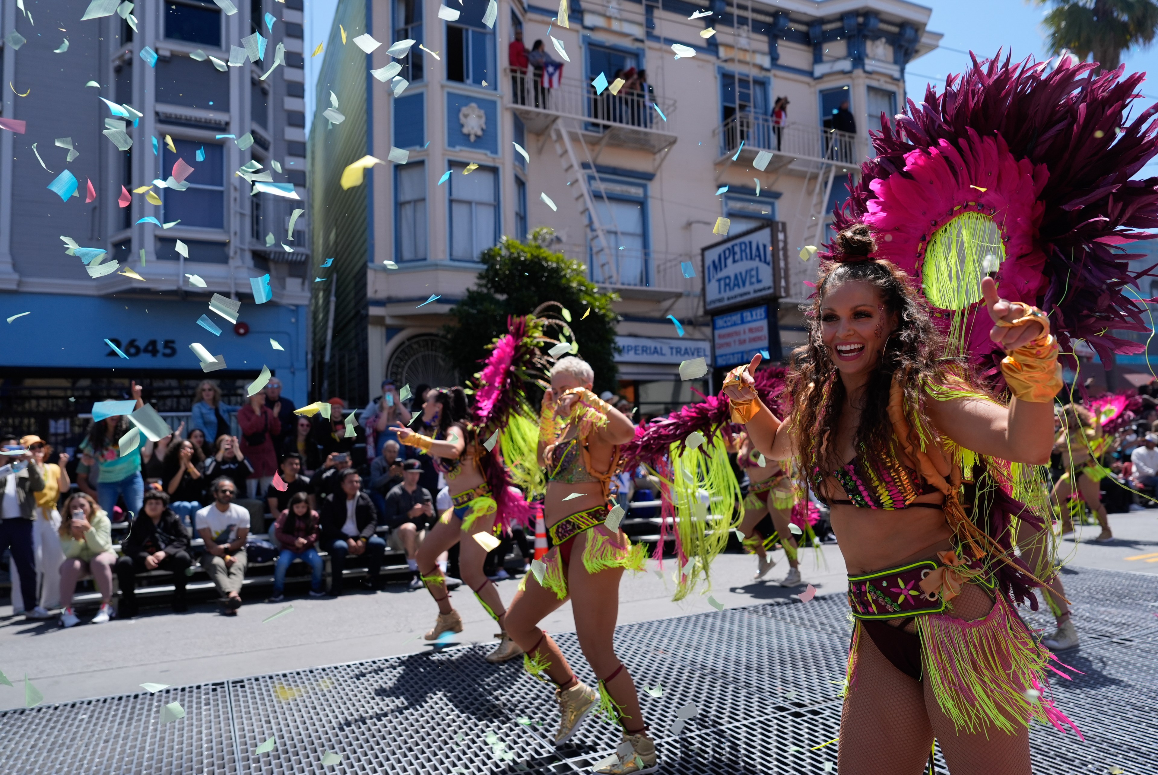 A vibrant street parade with dancers in colorful feathered costumes moves down the street, surrounded by confetti and cheering spectators.