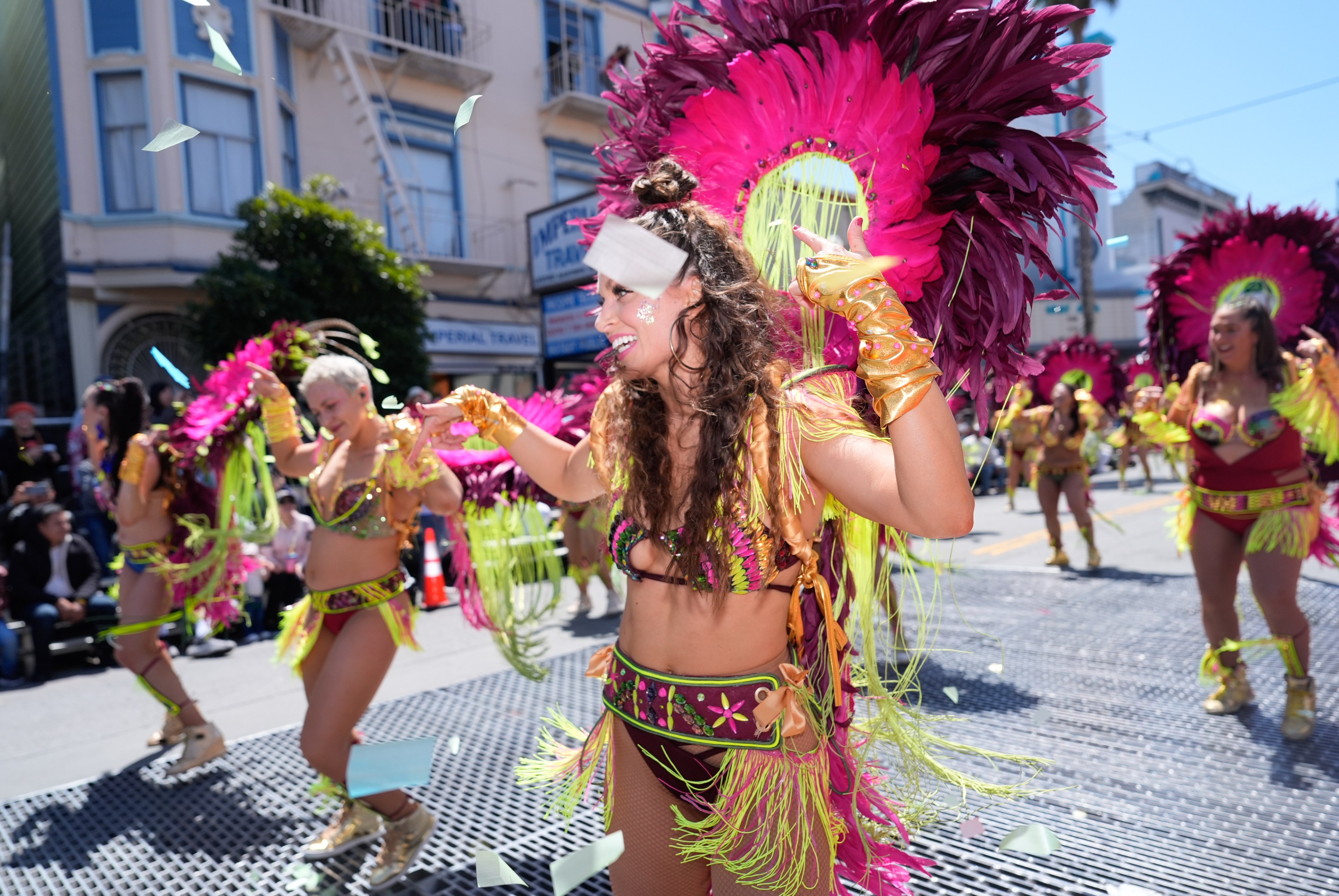 A vibrant parade scene features dancers in colorful, feathered costumes with pink and green accents, performing energetically on a city street.