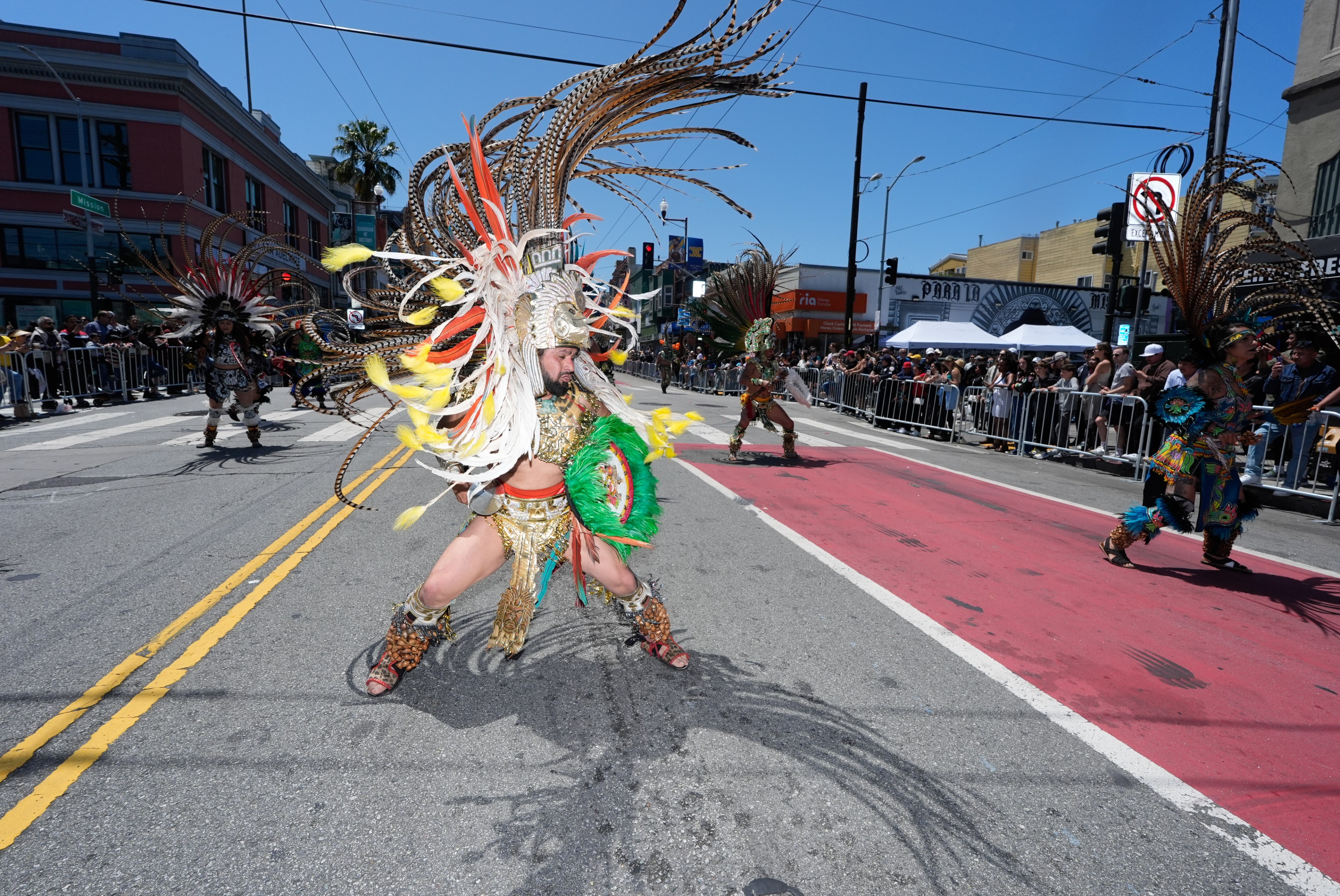 Dancers in vibrant traditional feathered costumes perform in a street parade, while onlookers stand behind barricades along the sidewalk on a sunny day.