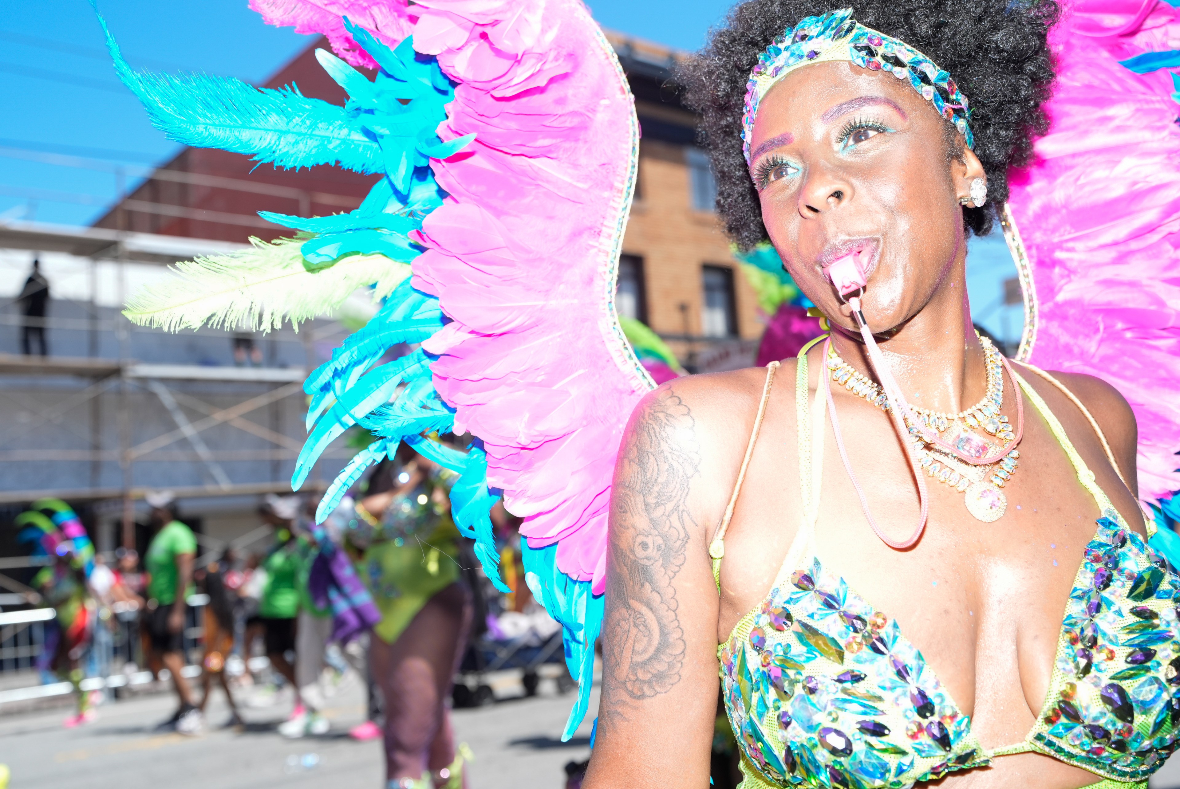 A person dressed in colorful carnival attire with large, vibrant pink and blue feathered wings is blowing a whistle. They have multiple necklaces and a sequined outfit.