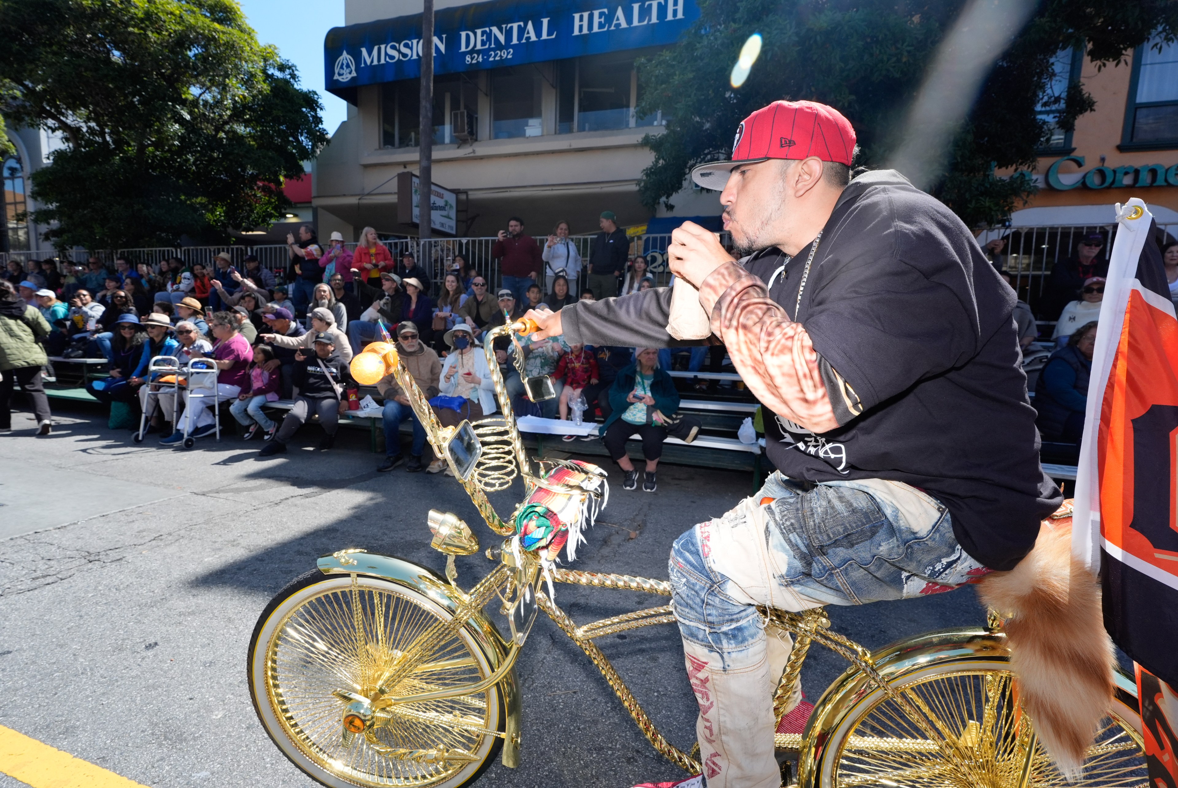 A man in a red cap and jeans rides a shiny gold bicycle past a seated crowd on city streets during a parade. The crowd watches attentively on a sunny day.