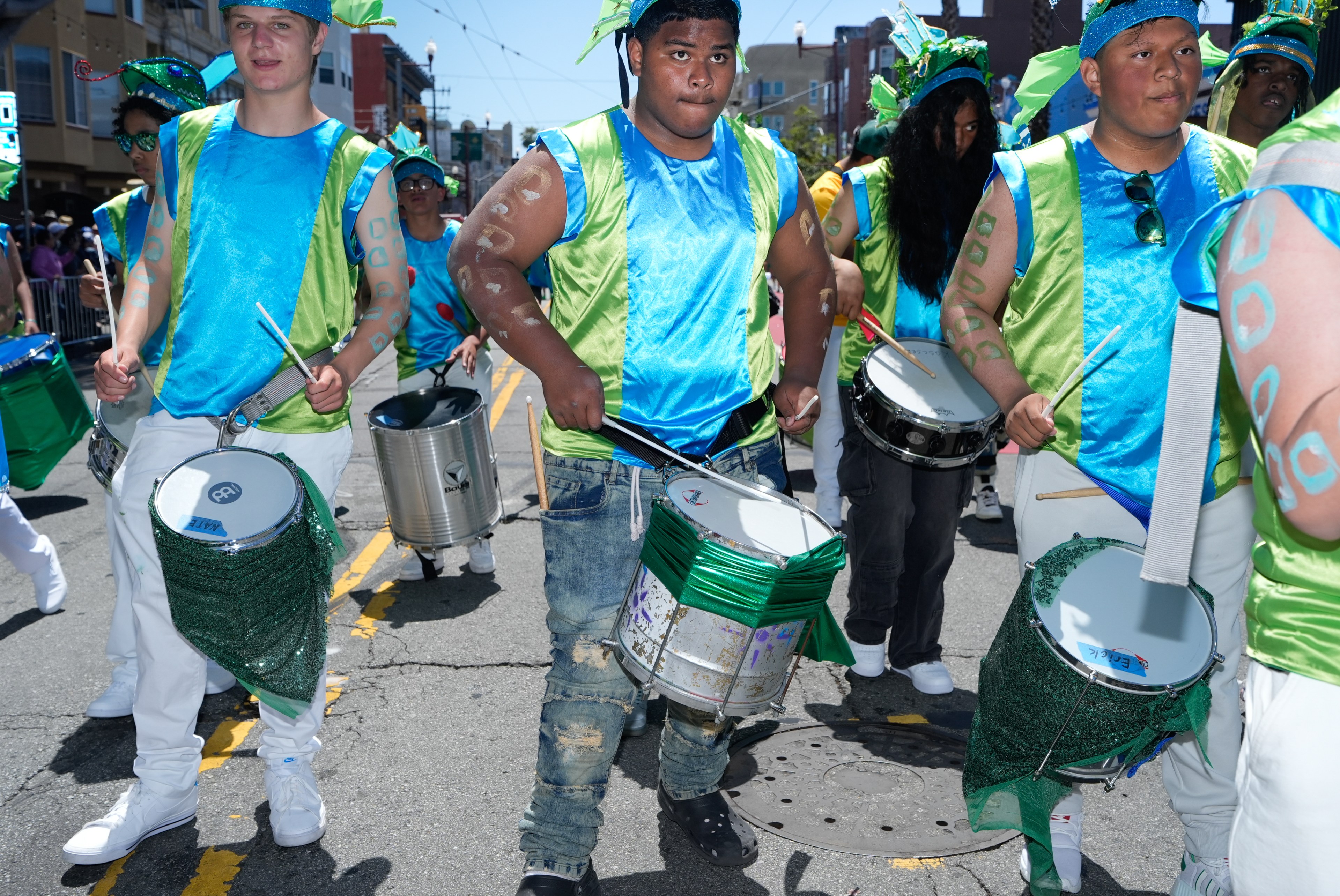 A group of people in vibrant blue and green outfits march down a street playing drums. They wear headgear and body paint, with a lively, festive atmosphere.