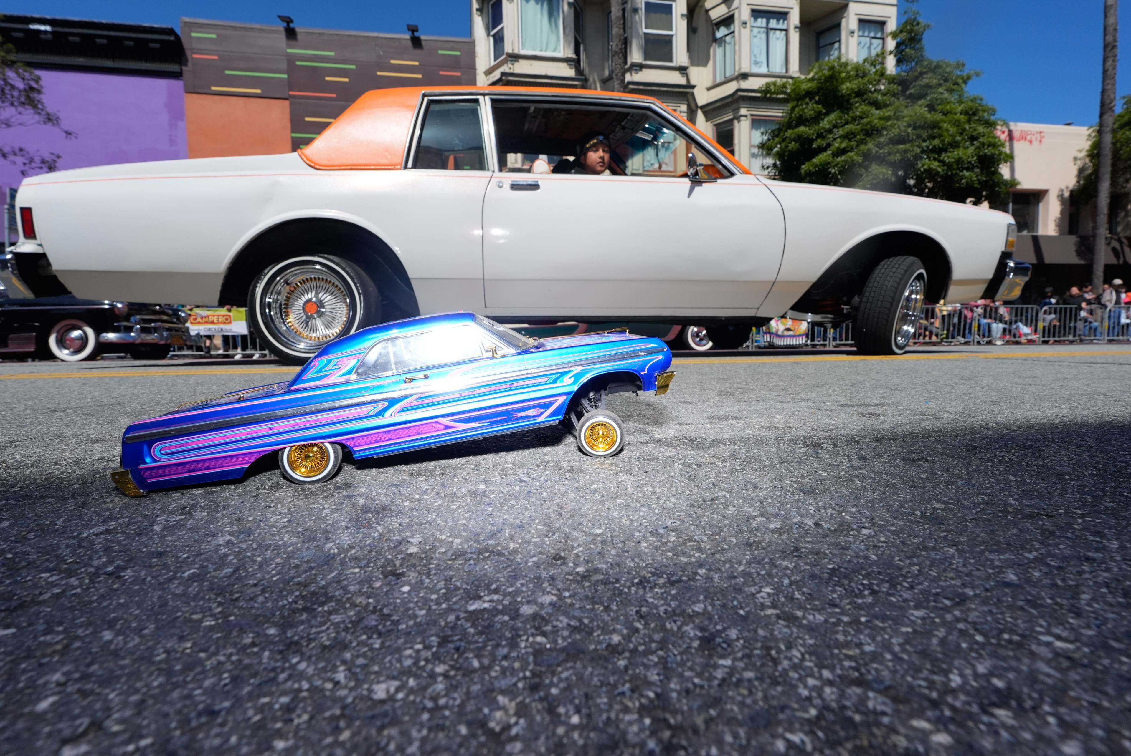 A small, colorful toy car with elaborate blue and purple designs is on the street, positioned in front of a larger white car with an orange roof and chrome wheels.