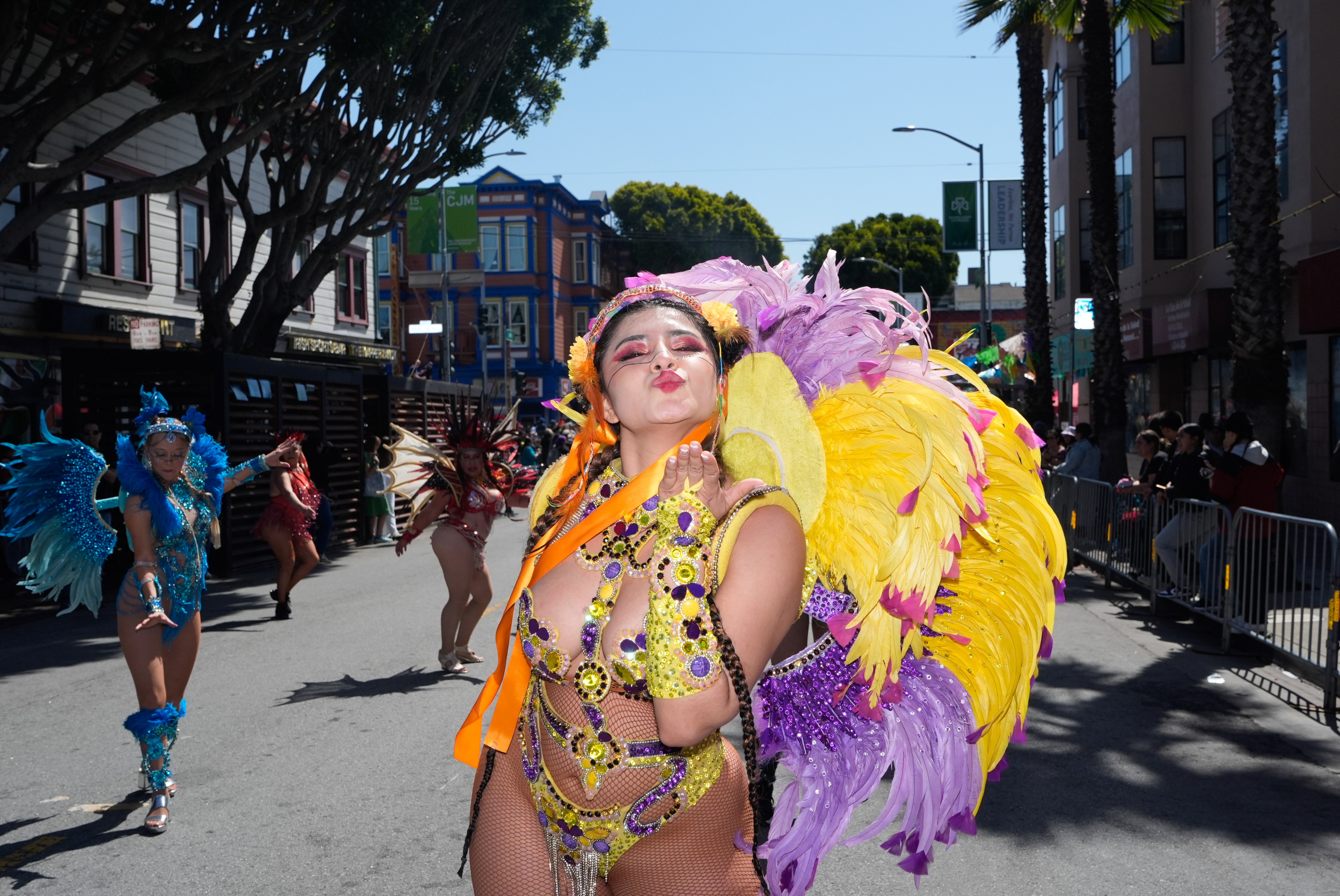A woman in colorful, feathered costume blows a kiss at a street parade. Others in vibrant outfits and large feathered headdresses dance in the background.
