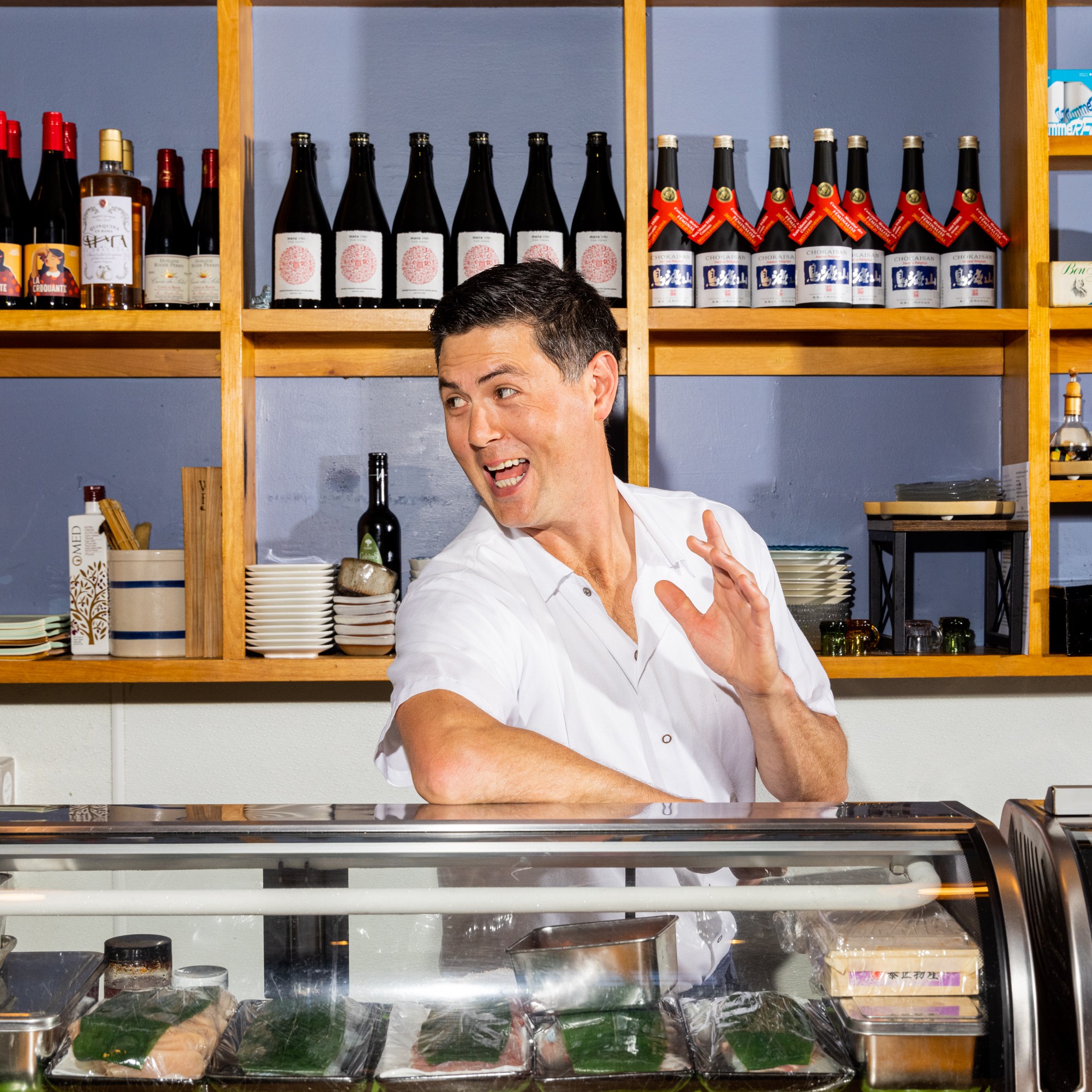 A man in a white shirt stands behind a counter in front of shelves stocked with bottles and dishes, smiling and gesturing with his right hand raised.