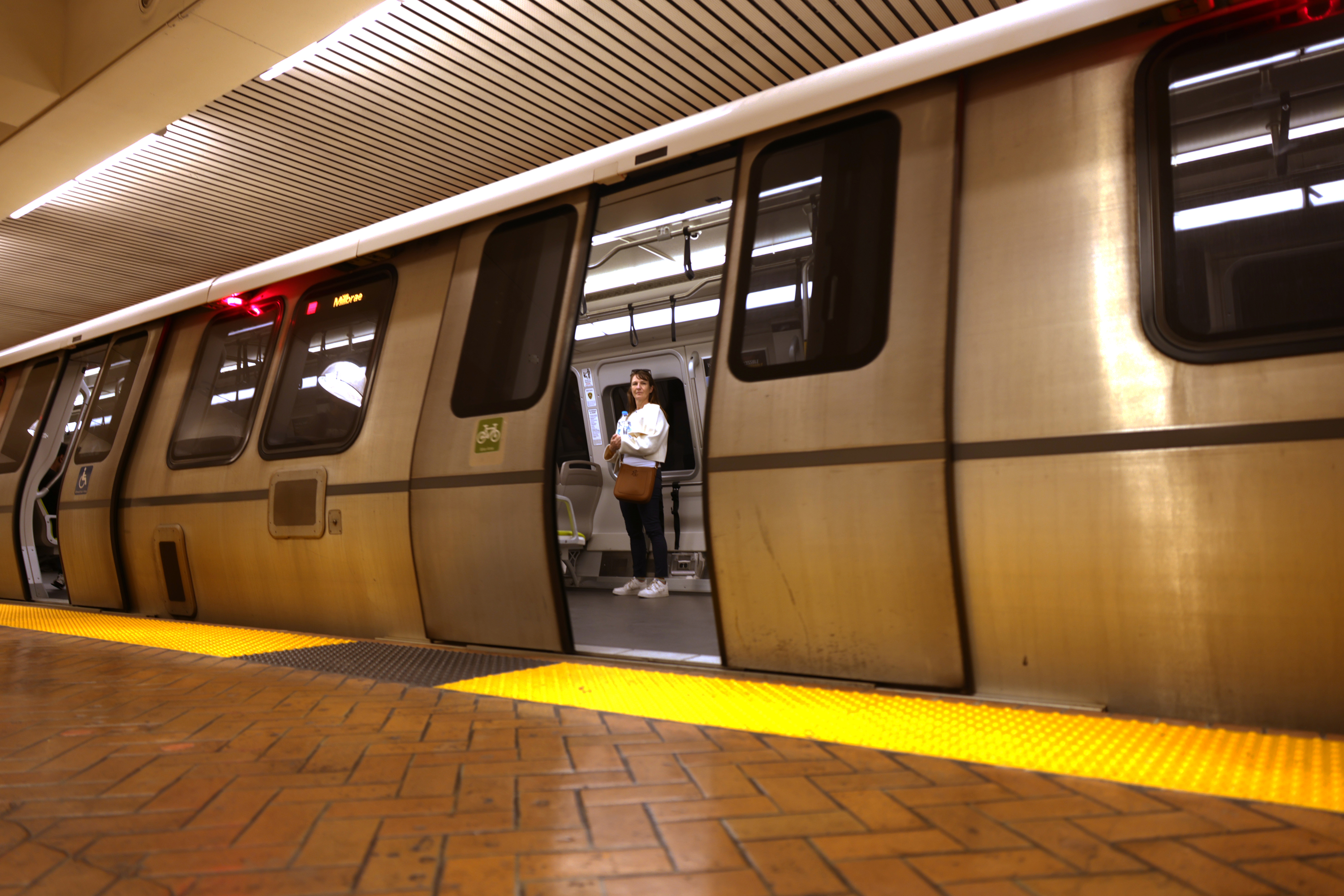 A lone person stands inside a stationary, open-doored subway car, with a beige interior, on a platform with yellow safety markings and brown tiles.