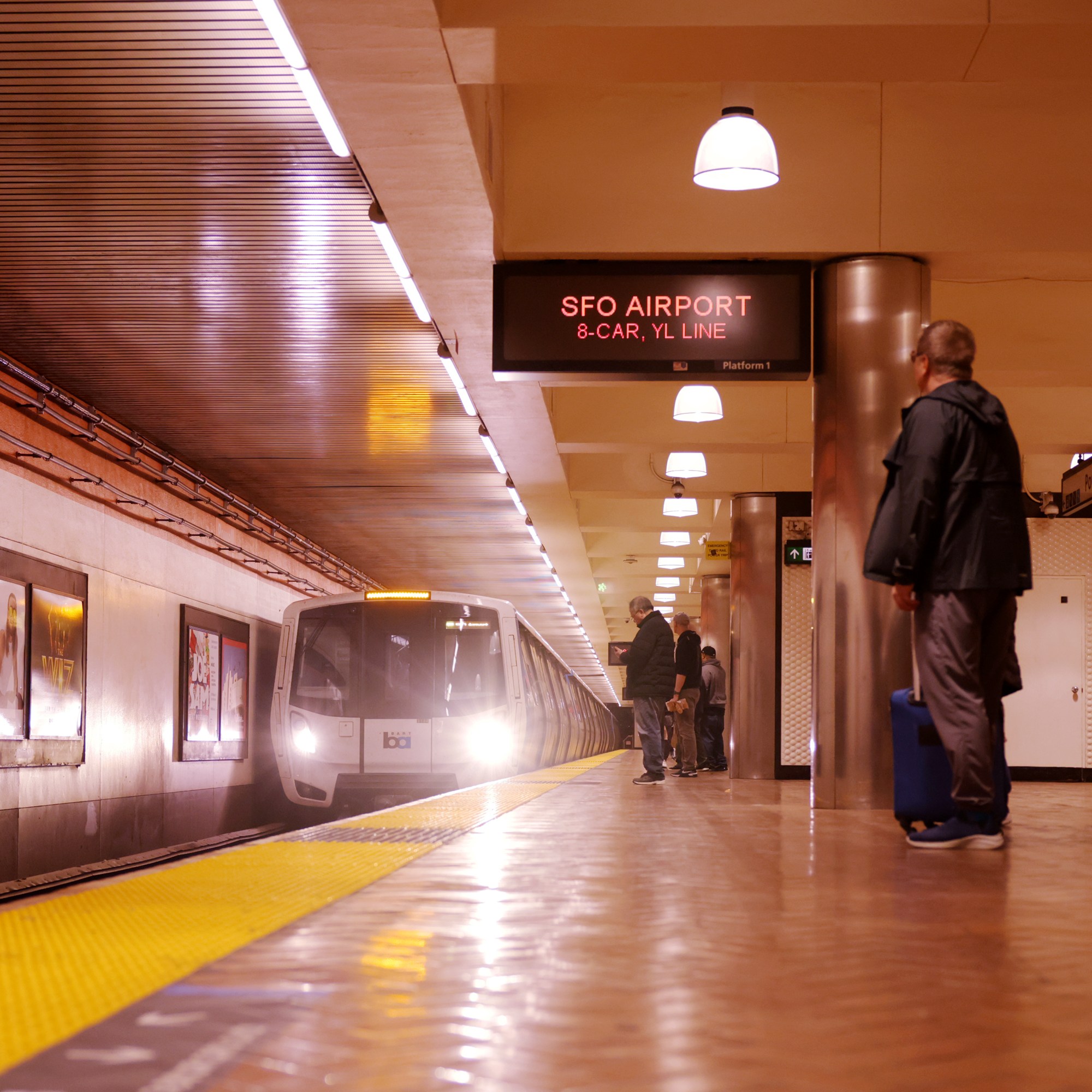 A subway platform with people waiting, a train approaching, and signs indicating the destination as "SFO Airport" on an 8-car Yellow Line.