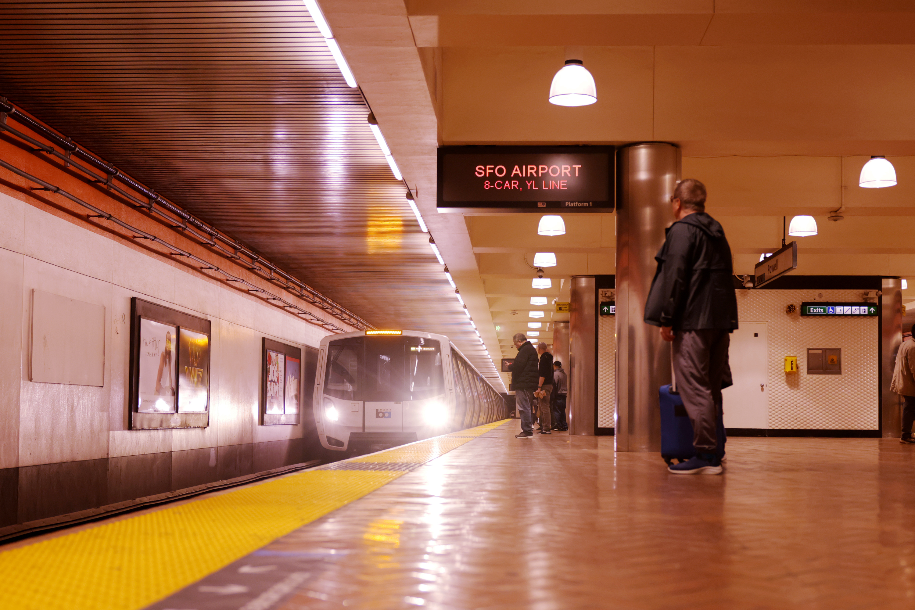 A subway platform with people waiting, a train approaching, and signs indicating the destination as "SFO Airport" on an 8-car Yellow Line.