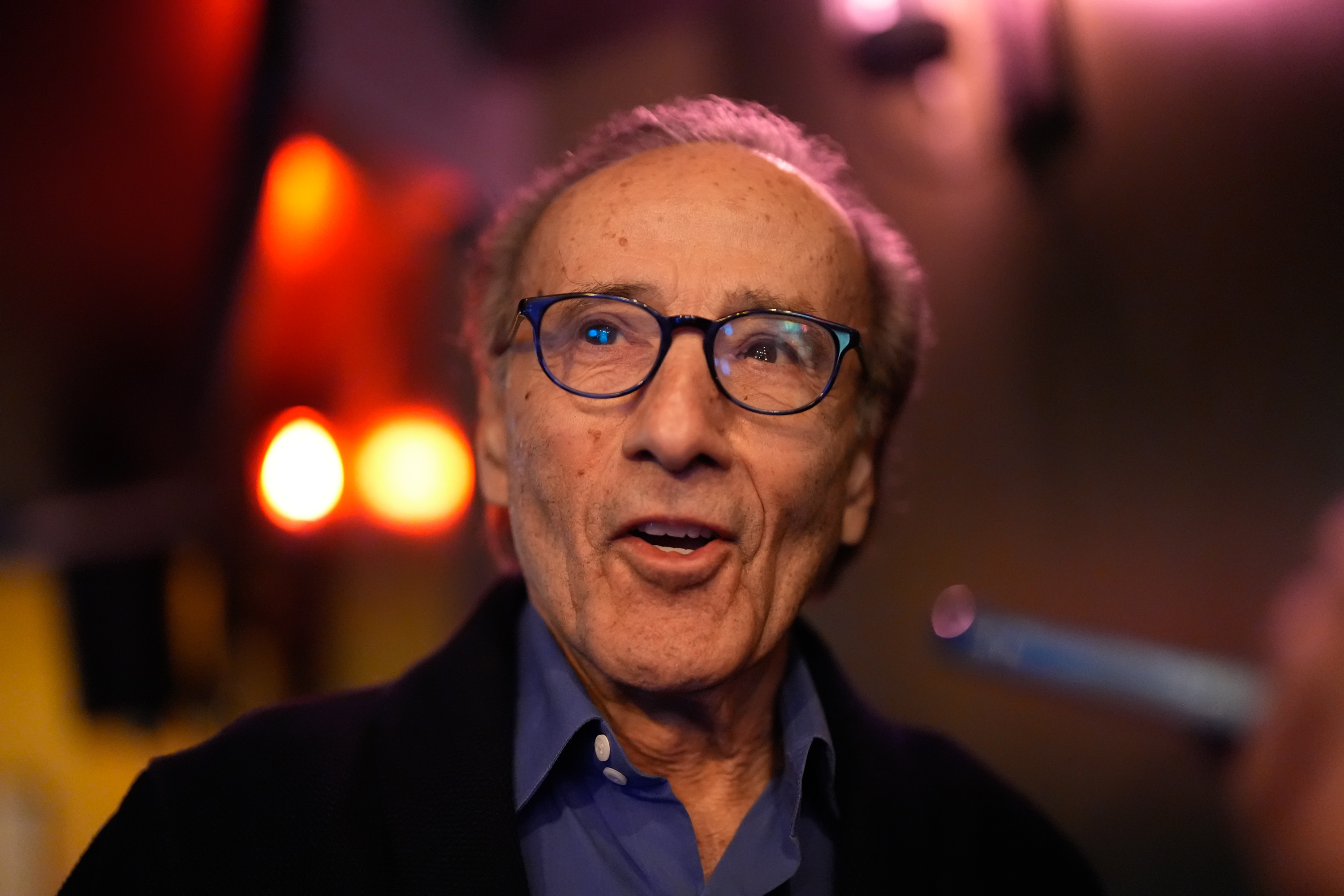 An elderly man with glasses and thinning hair is smiling in a dimly lit room with warm lights in the background.