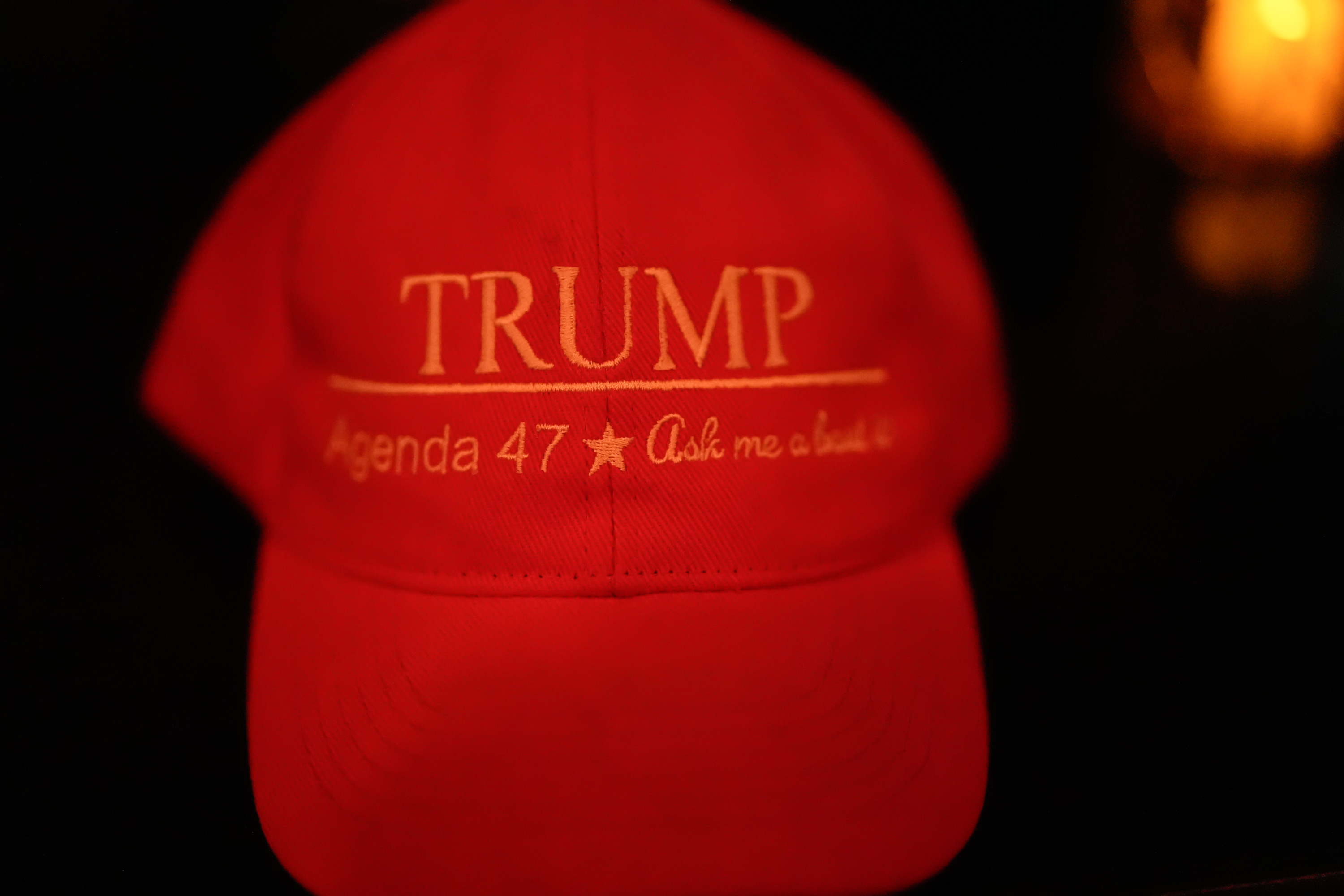 The image shows a red baseball cap with &quot;TRUMP&quot; in large letters and &quot;Agenda 47 - Ask me about it&quot; underneath. The background is dark.