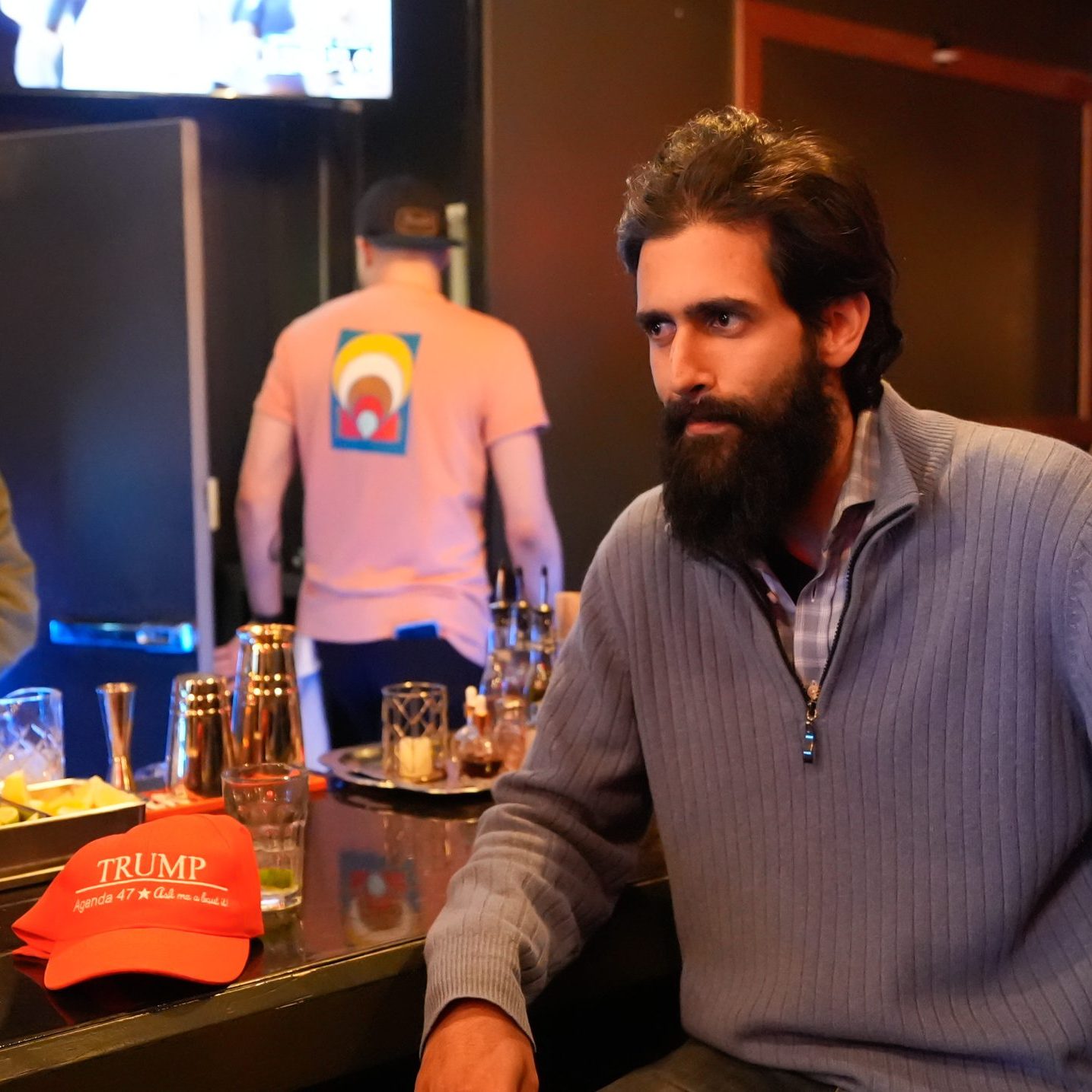 A man with a beard in a gray sweater is sitting at a bar counter, next to a red "Trump" hat. A bartender is preparing drinks and a TV is on in the background.