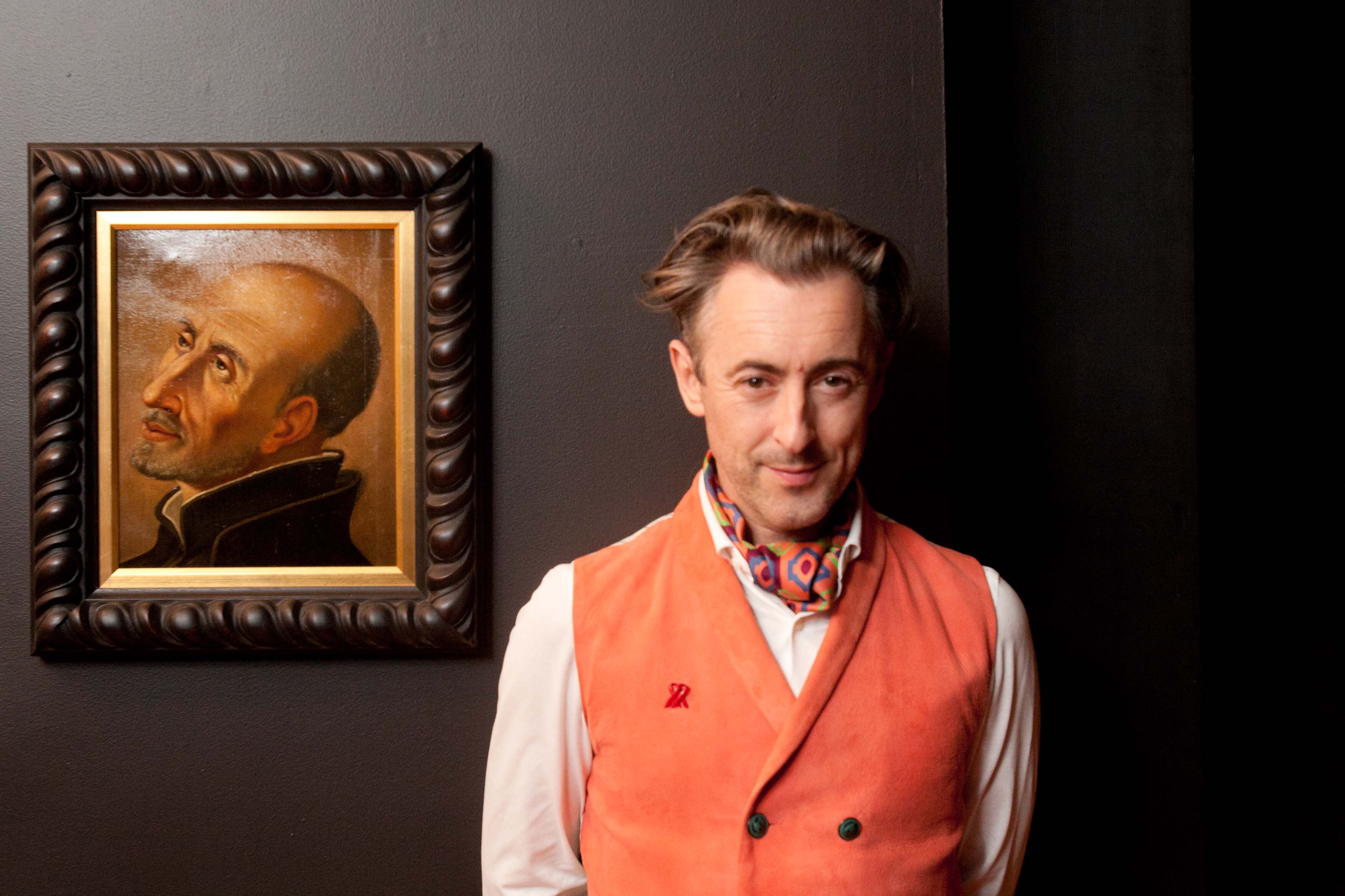 A person in an orange vest and colorful scarf stands next to a framed painting of a bald man with a contemplative expression, set against a dark background.