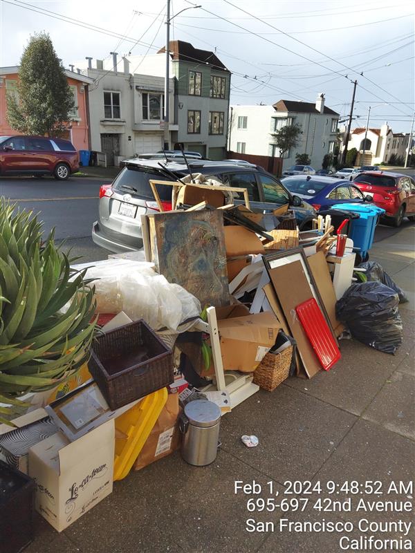 A pile of discarded items cluttering a sidewalk, with townhouses and parked cars in the background.