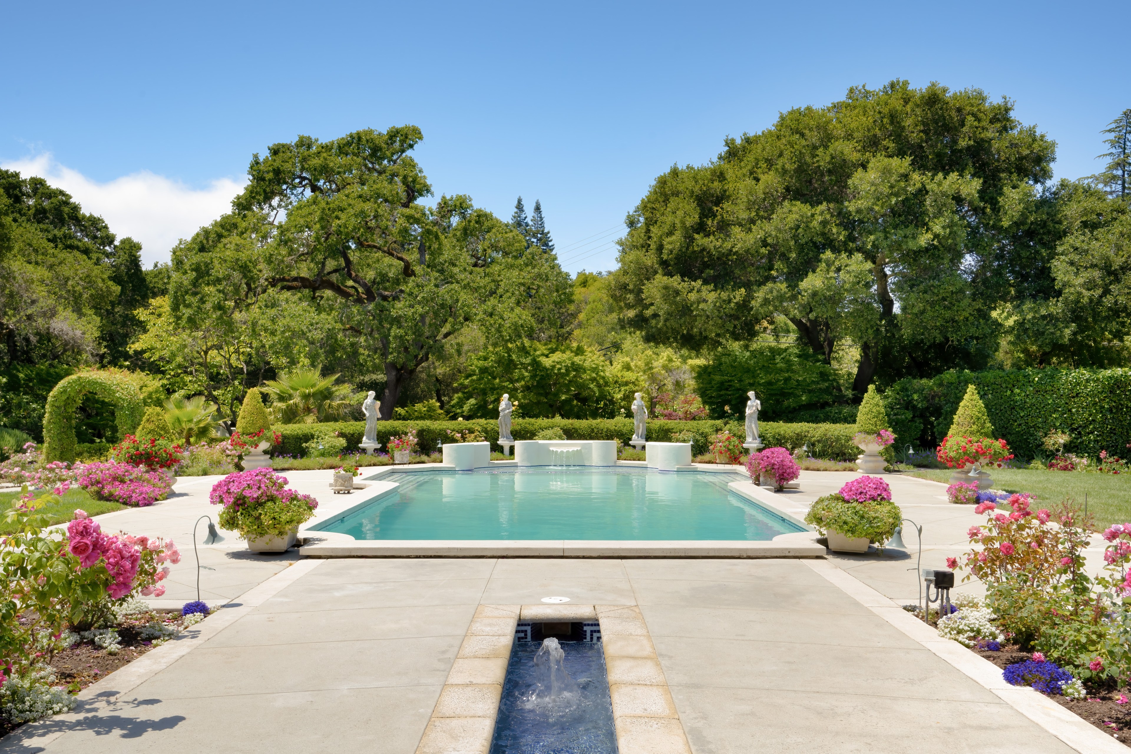 A swimming pool shimmers in sunlight next to flowers, a small fountain and a line of trees