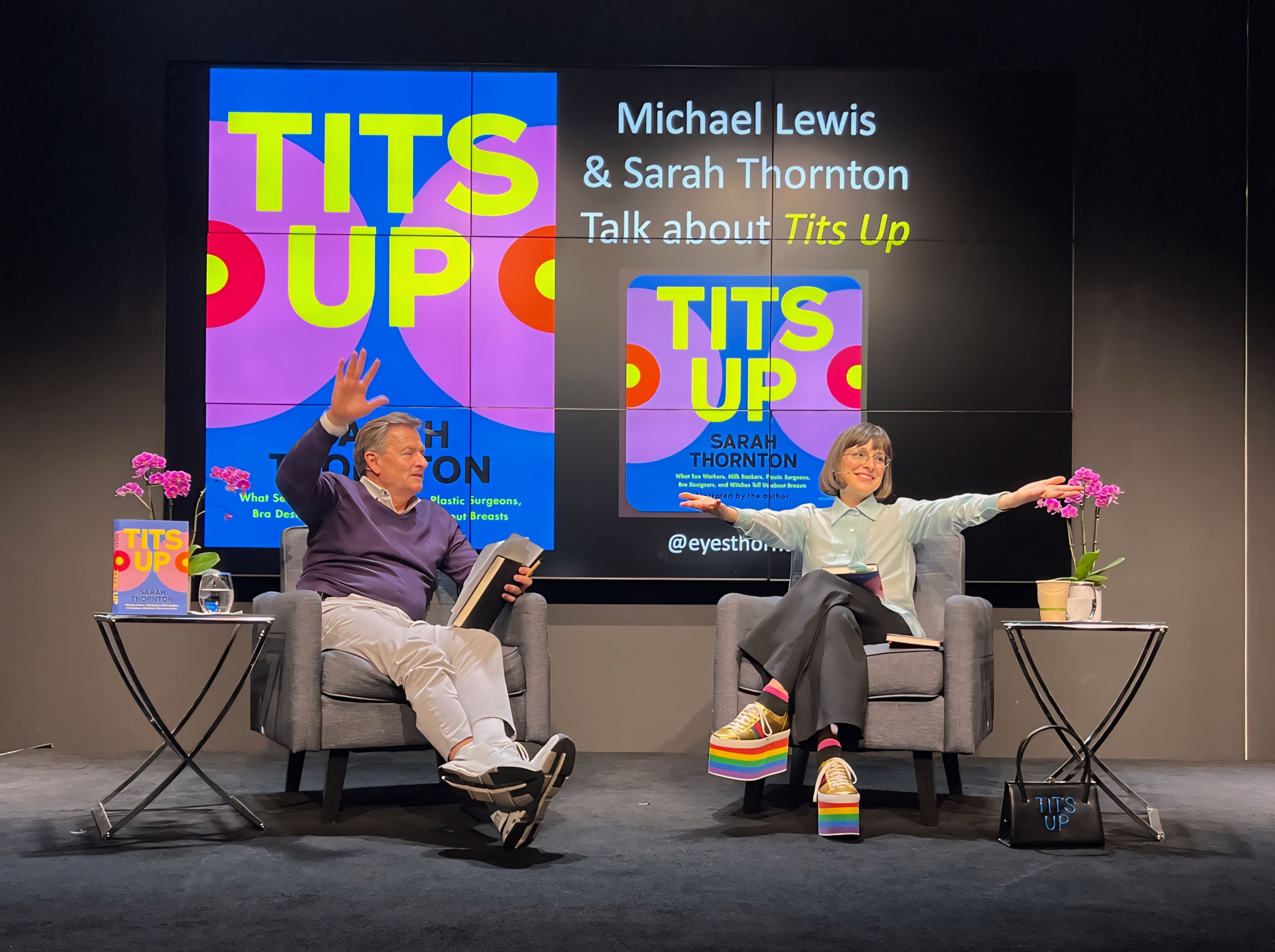 Two people are seated on stage waving, with a backdrop advertising a &quot;Tits Up&quot; talk and book.