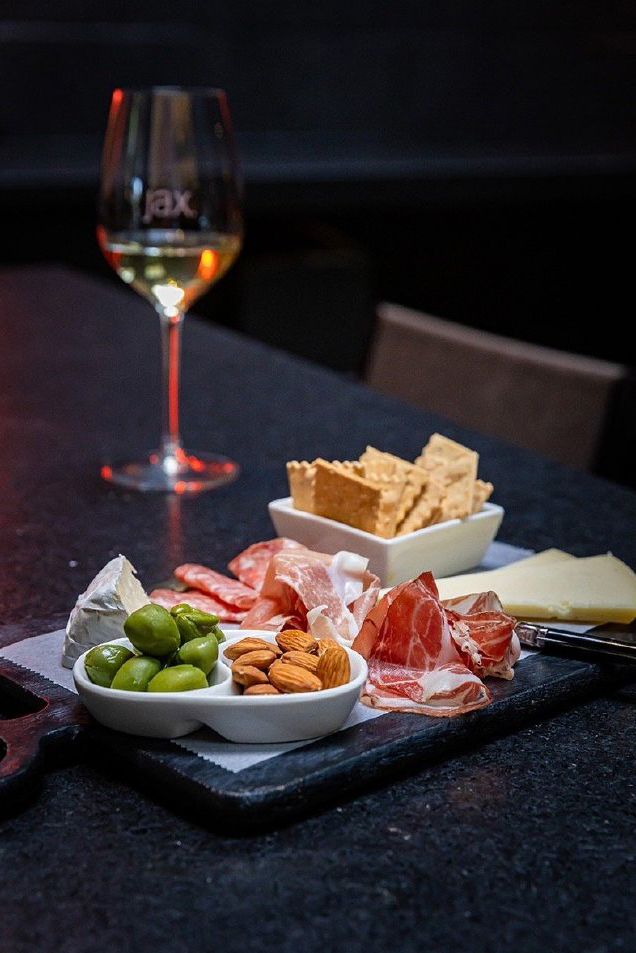 A glass of white wine next to a charcuterie board featuring various cheeses, prosciutto, almonds, and olives on a dark table.