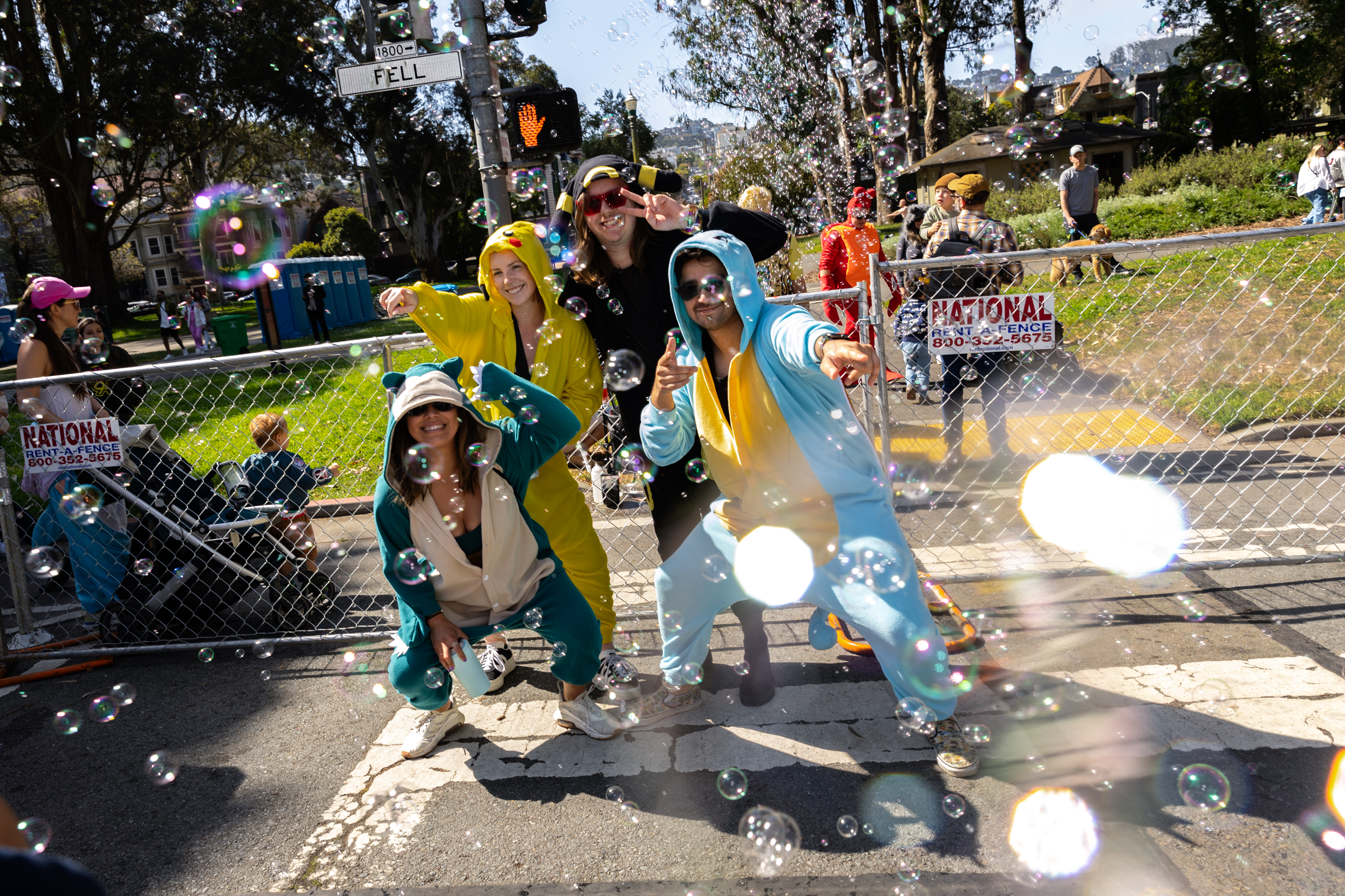 Four joyful people in animal onesies play with bubbles on a sunny street corner, surrounded by colorful floating bubbles and greenery.