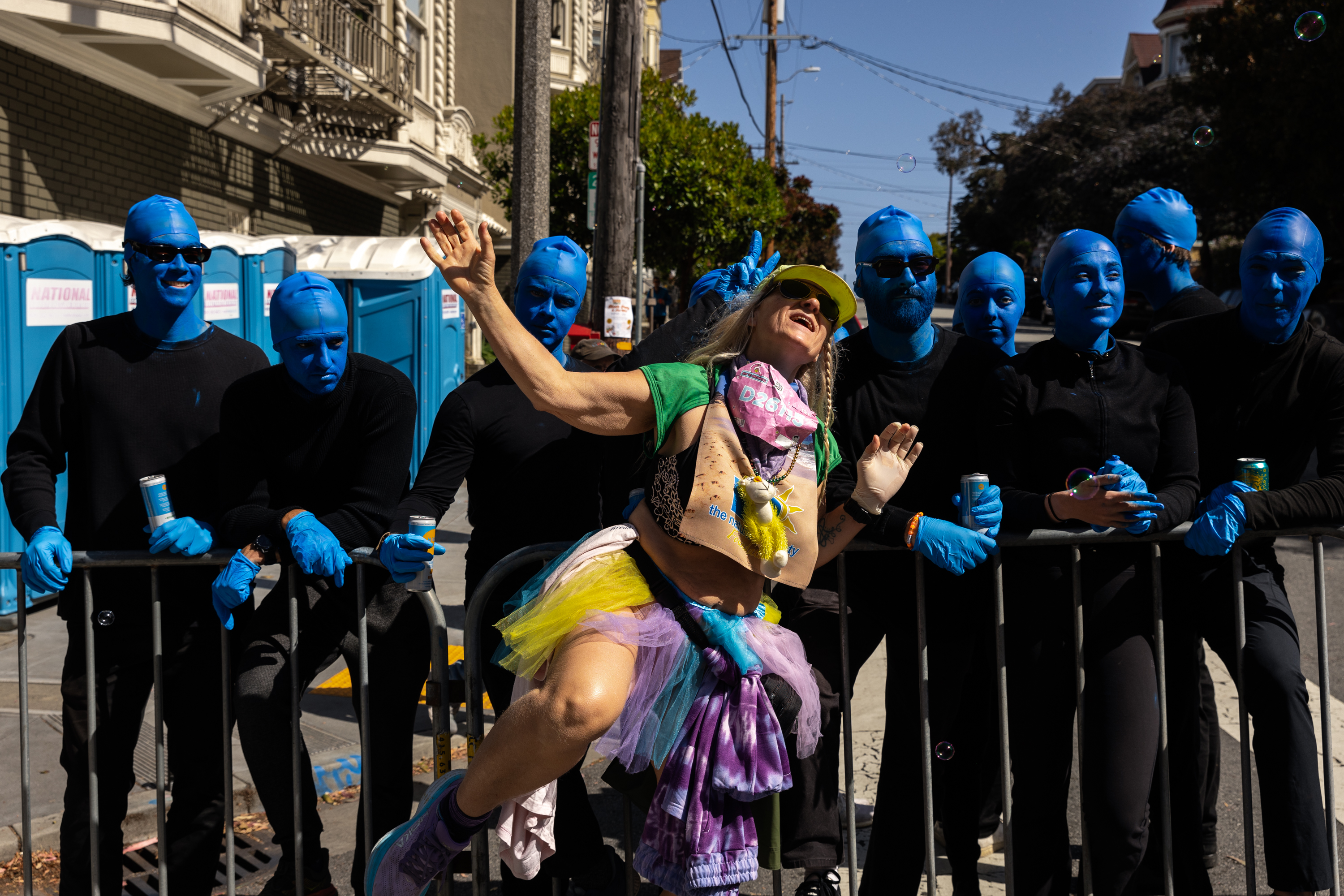 A woman in a colorful tutu dances jubilantly near blue-painted performers resembling Blue Man Group, who are behind a barricade.