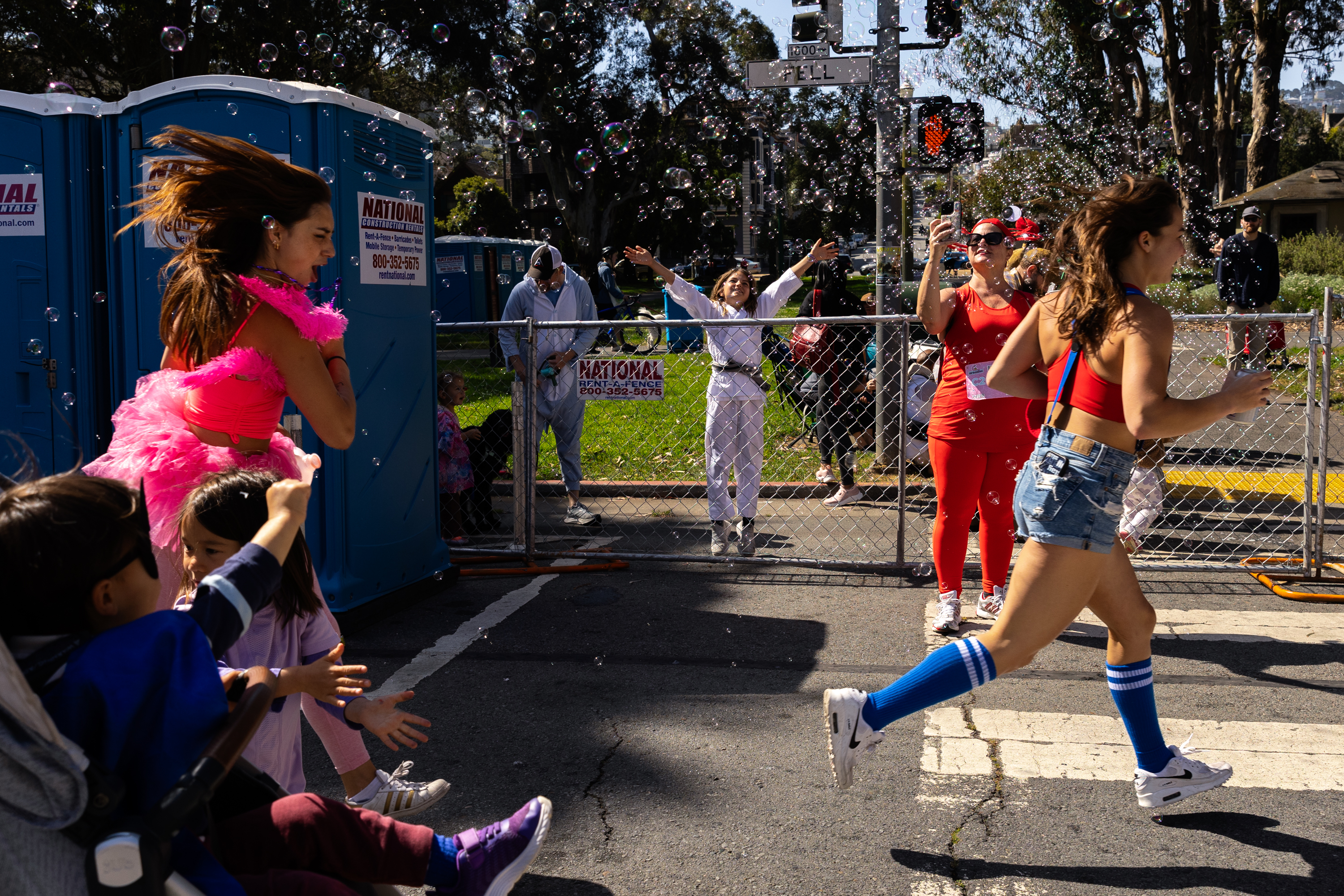 A lively street scene with people dressed in colorful, festive outfits, including a woman in pink, running and generating bubbles, with spectators, including children, enjoying the scene.