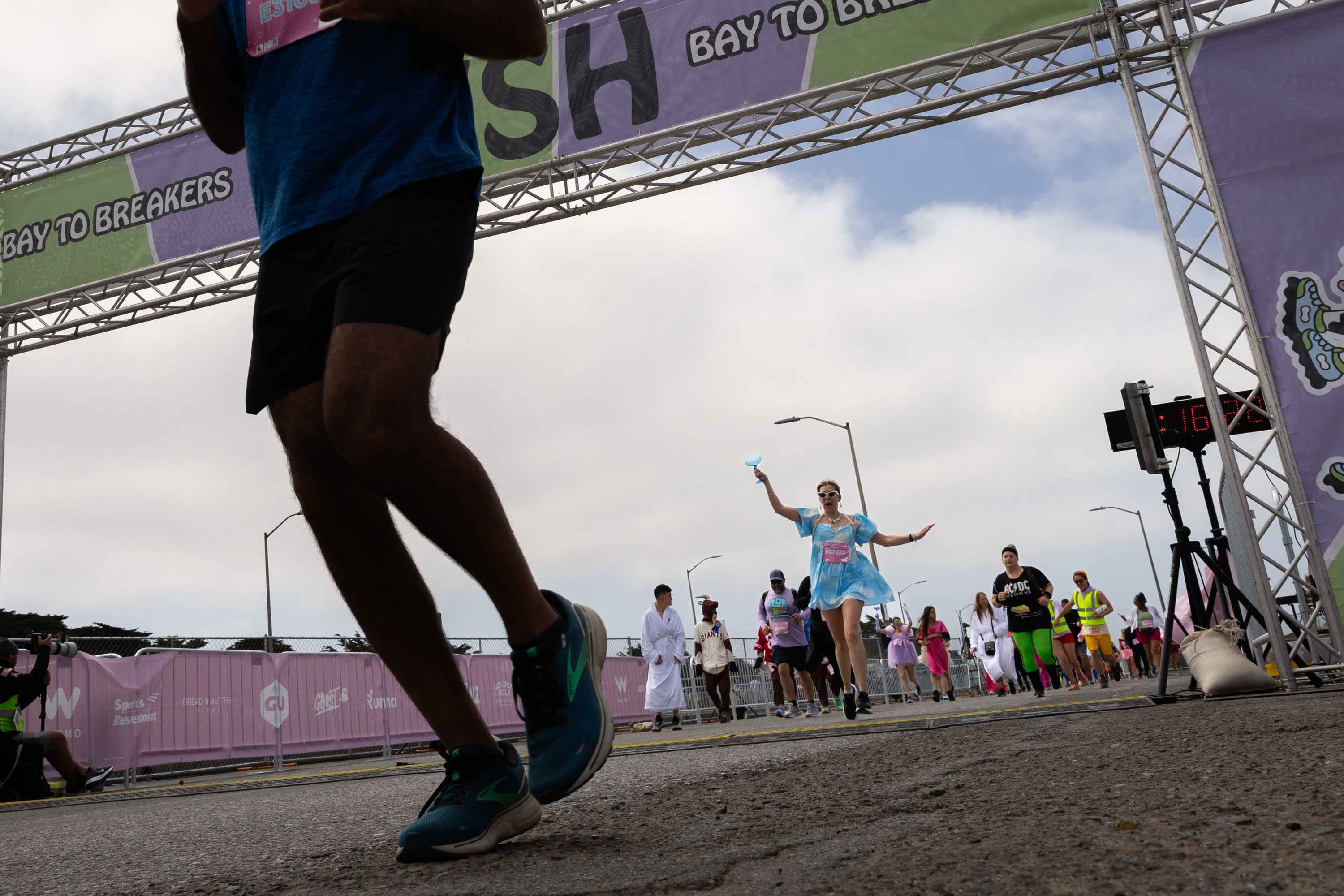 Runners cross a finish line under a banner reading &quot;Bay to Breakers&quot;, including one woman in a light blue tutu, arms spread wide in celebration.