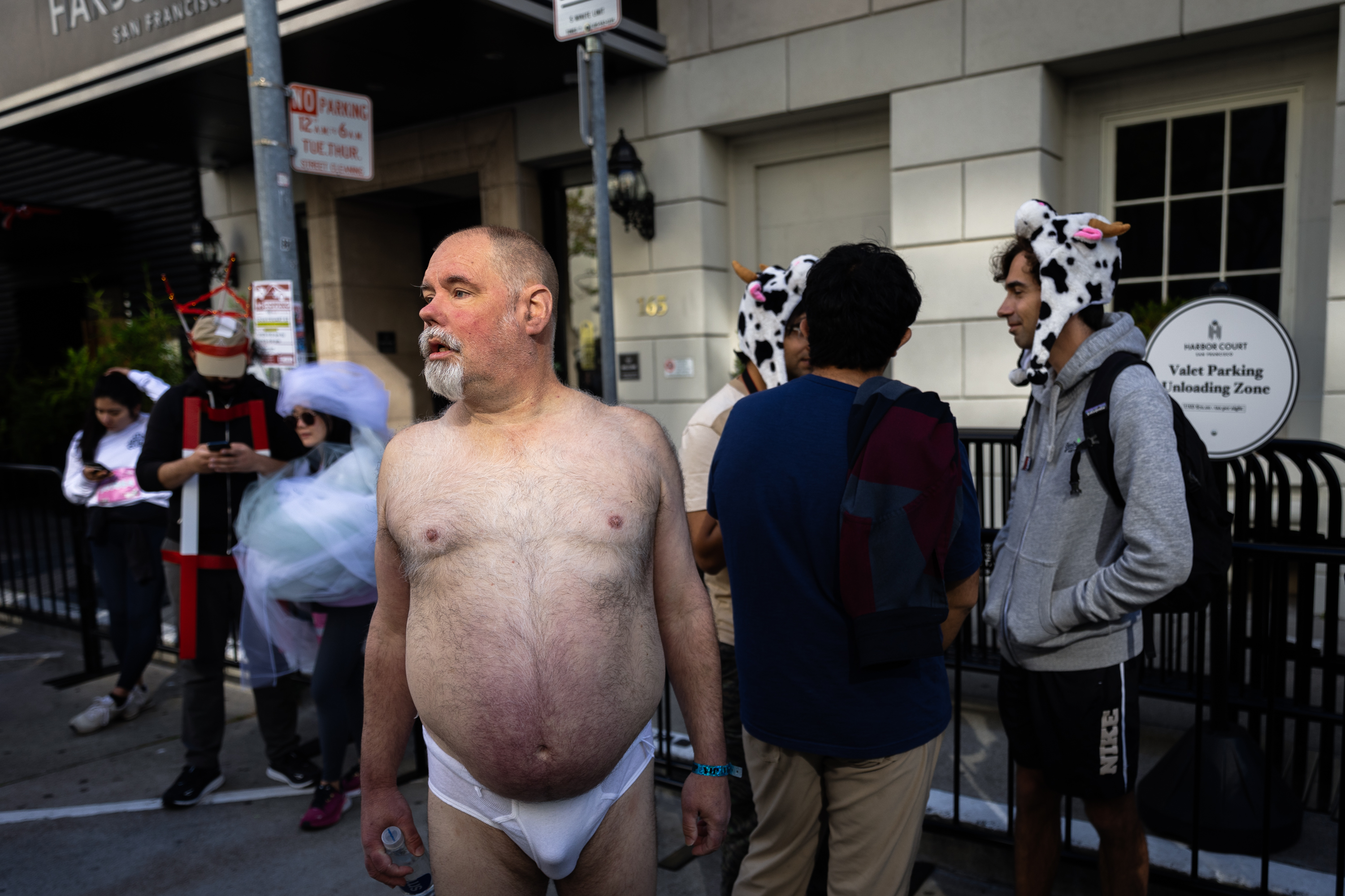 A man without a shirt, wearing only a white brief, stands in a city street crowded with festively dressed people.